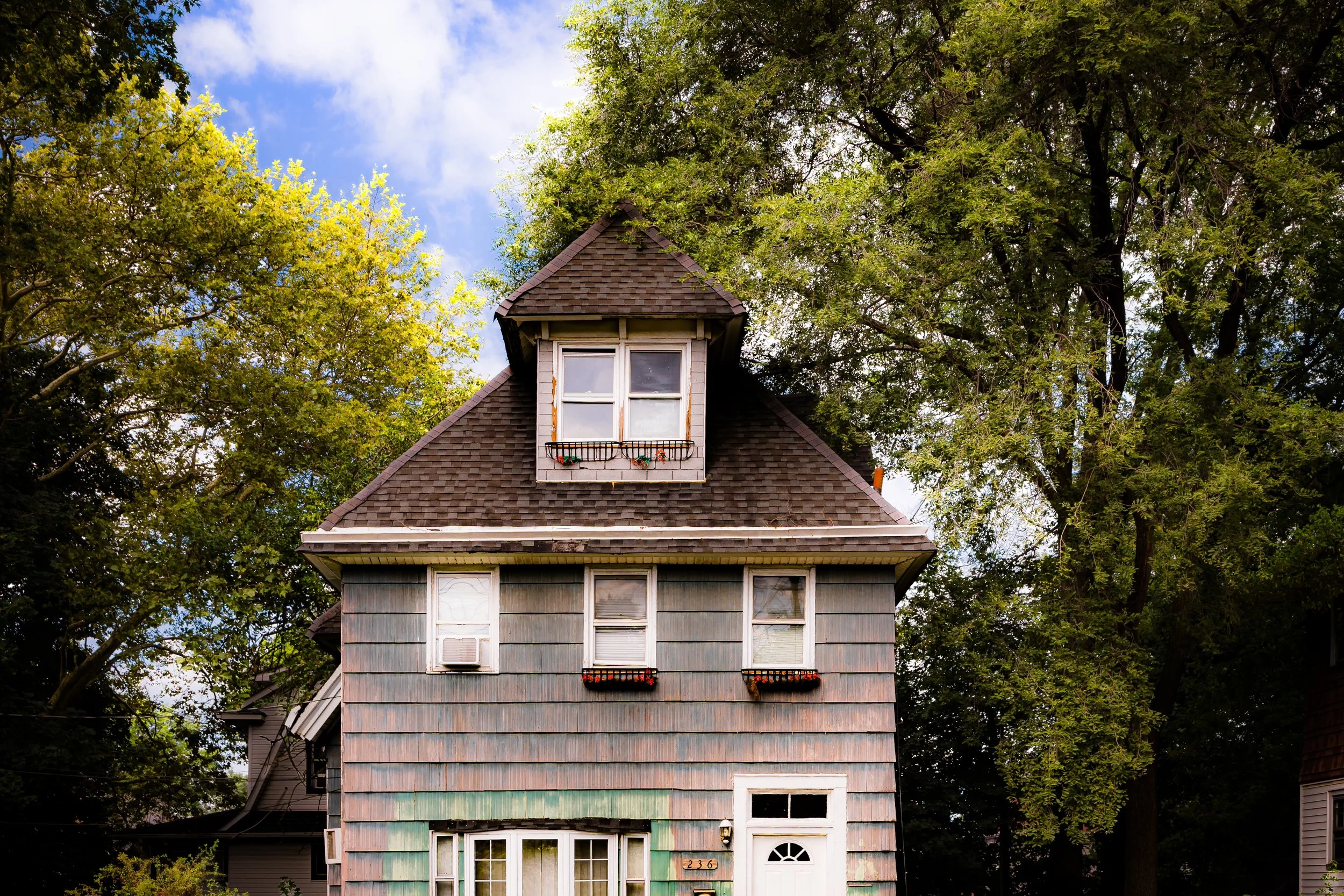 Old two-story house with weathered gray siding and a steep roof, surrounded by large green trees and a partly cloudy blue sky.