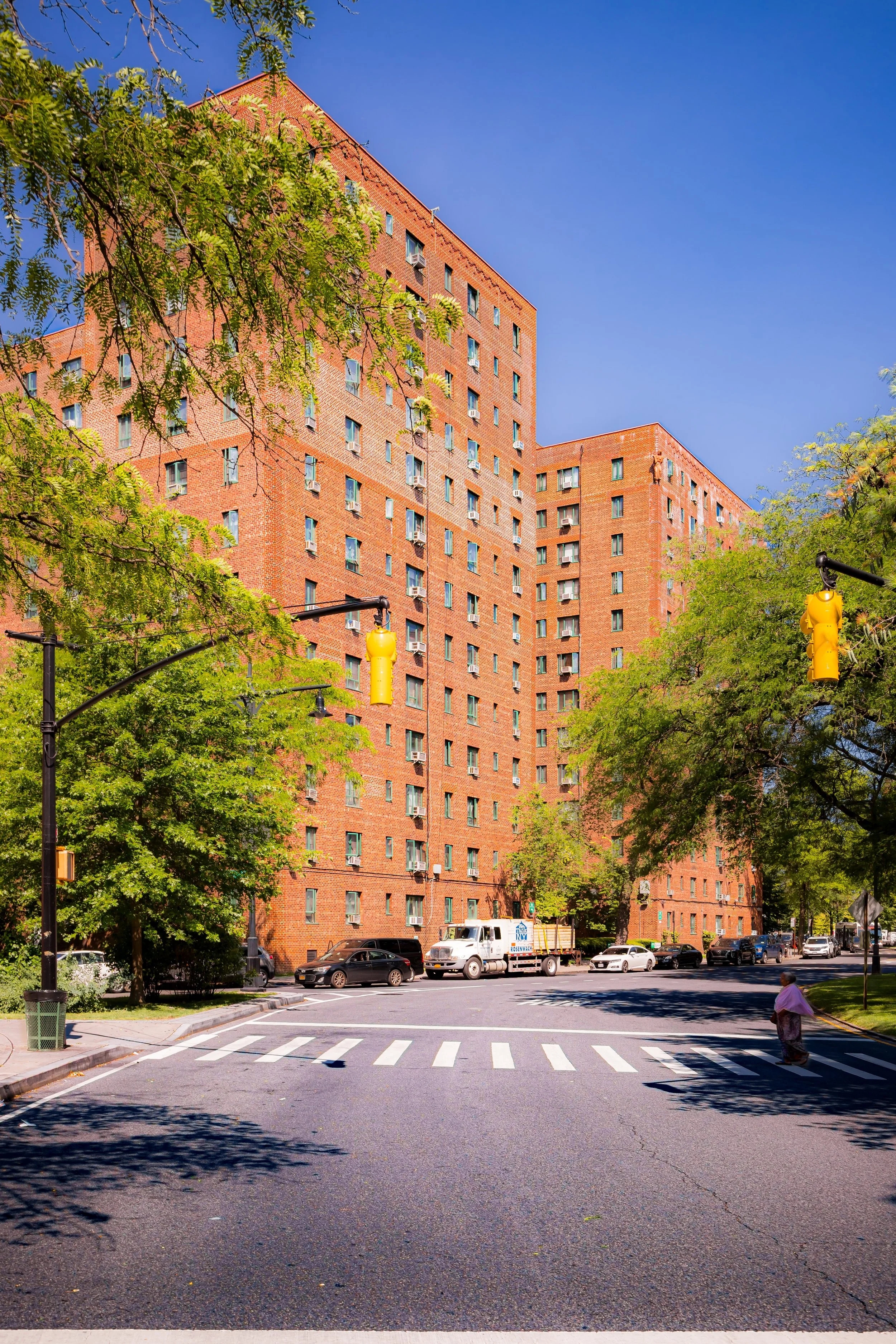 A tall red brick apartment building seen from across the street with a crosswalk in the foreground, trees on either side, and parked cars along the curb under a clear blue sky.