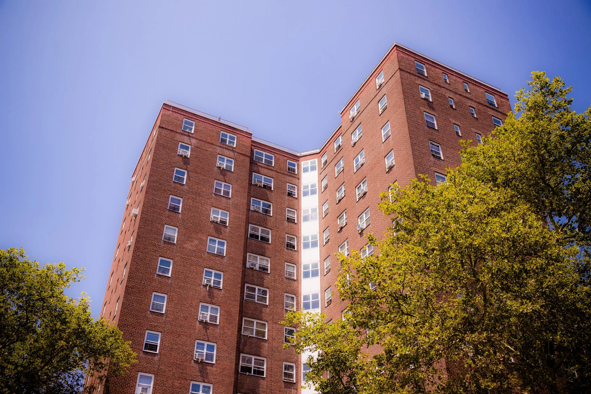 A tall brick apartment building viewed from the ground looking up, with a clear blue sky and green trees in the foreground.