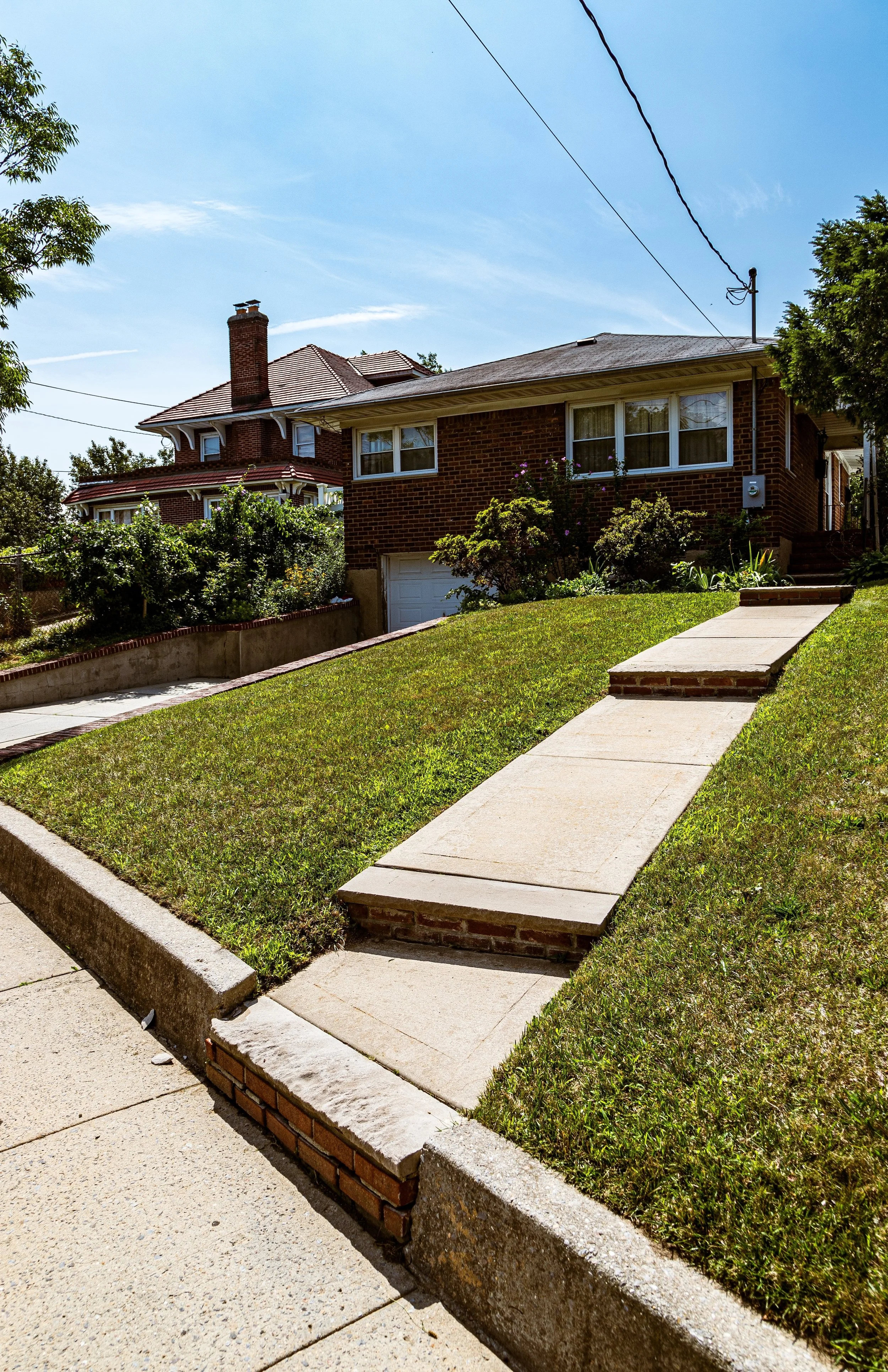 Front yard with a sloped grassy lawn, a walkway, and a brick house with large windows in the background.