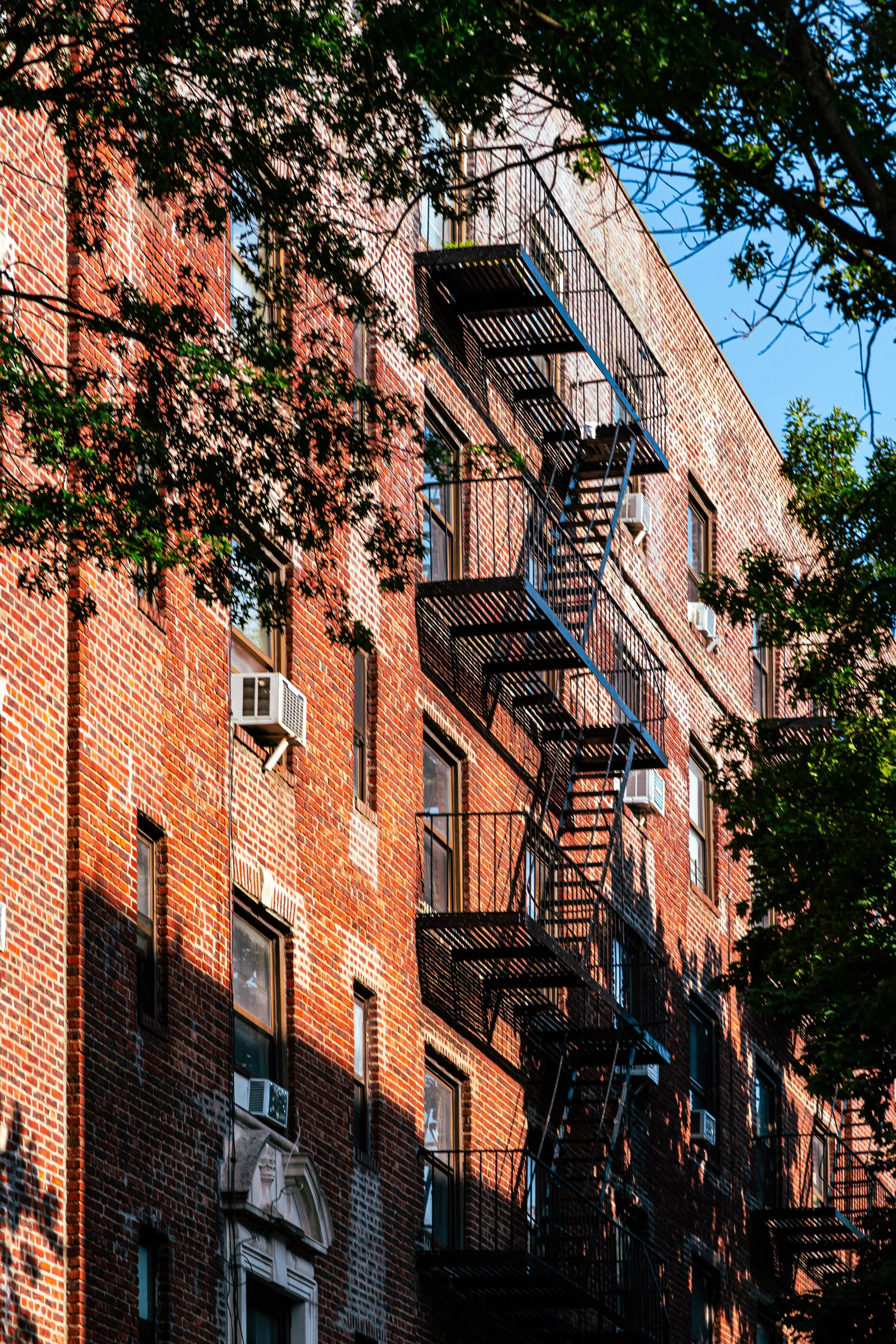 A brick apartment building with multiple fire escape staircases on the front. Trees partially obscure the building, and the sky is clear and blue.