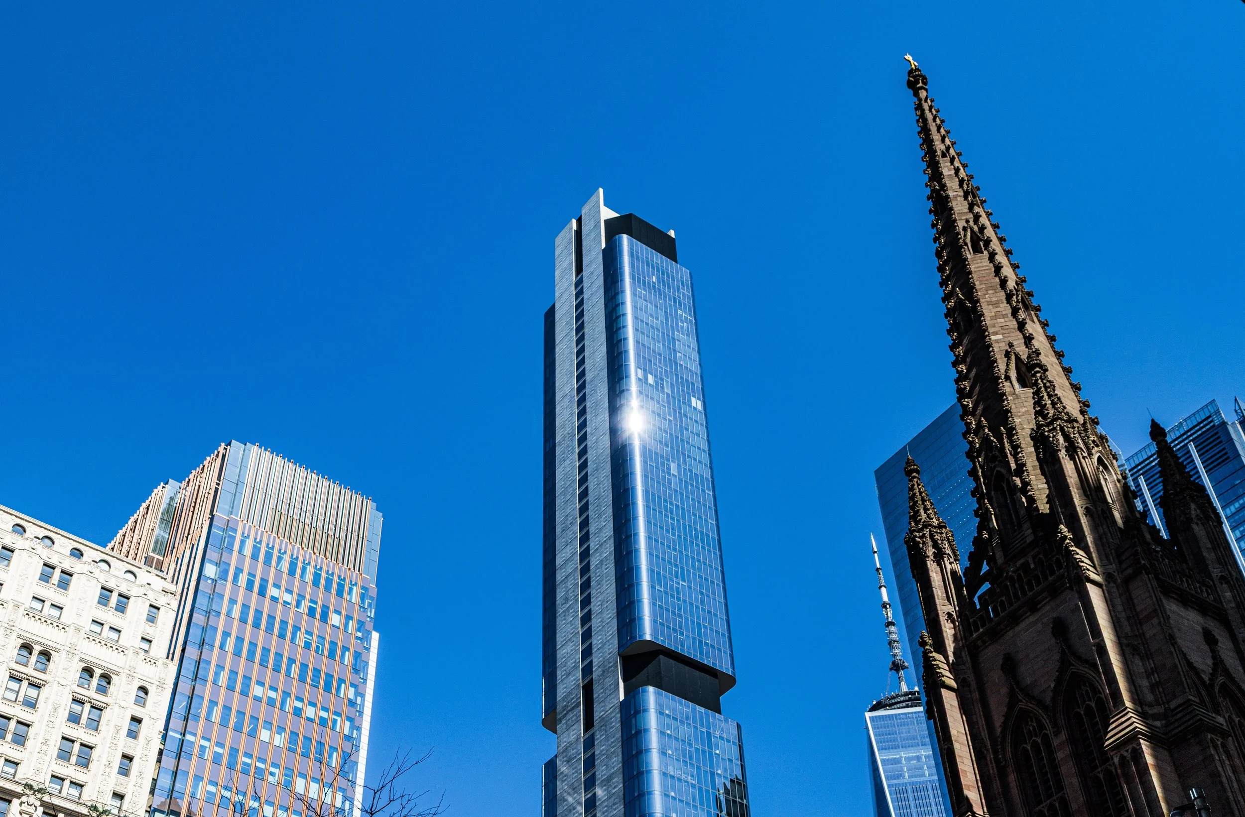 A cityscape featuring tall modern skyscrapers, a historic church with gothic architecture, and the Empire State Building in the background, all under a clear blue sky.