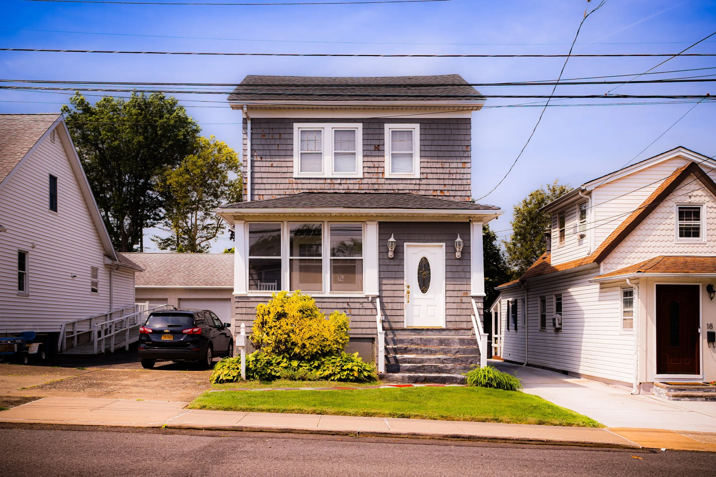 A two-story house with gray wooden shingles, white trim, and a white front door with decorative glass. The house has a front porch with steps, two lantern-style lights, and a small garden shrub in front. To the left, there is a black car parked in a driveway, and neighboring houses are visible on both sides. Overhead, utility wires are seen against a partly cloudy sky.