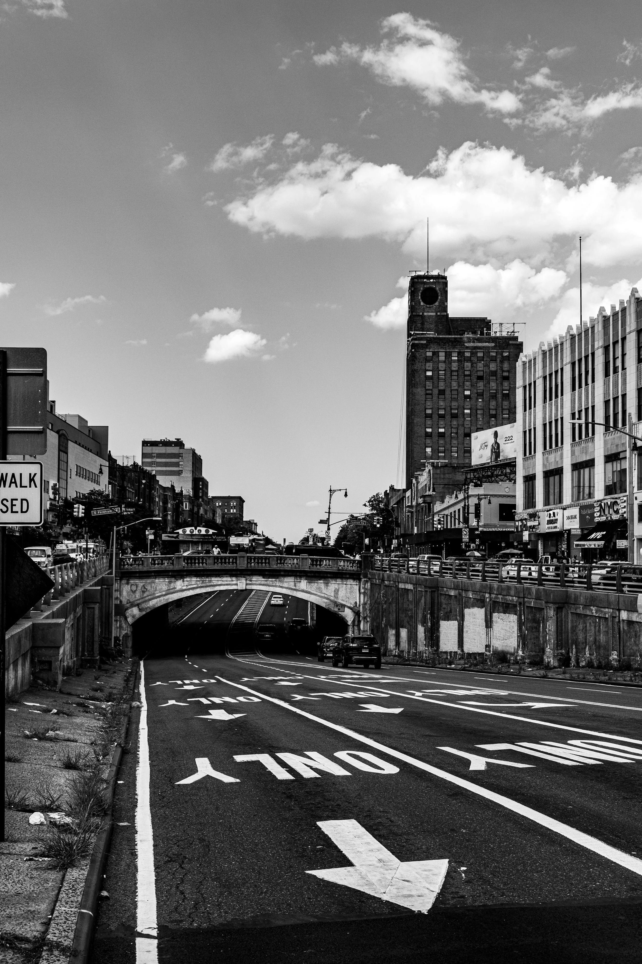 Black and white urban street scene with a road leading under a bridge, tall buildings, parked cars, and a cloudy sky.
