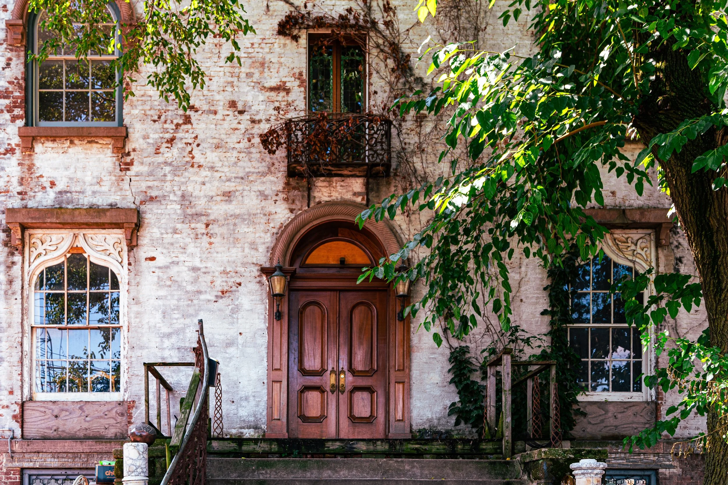 An old brick house with large windows, decorative wood framing, greenery around the entrance, a wooden door, and a small balcony with a wrought iron railing.