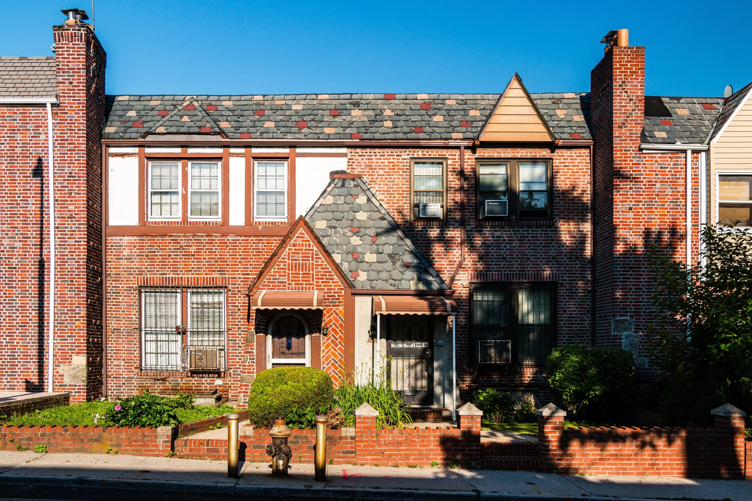 A row of red brick townhouses under a blue sky, with front gardens and small fences; shadows cast by nearby trees.