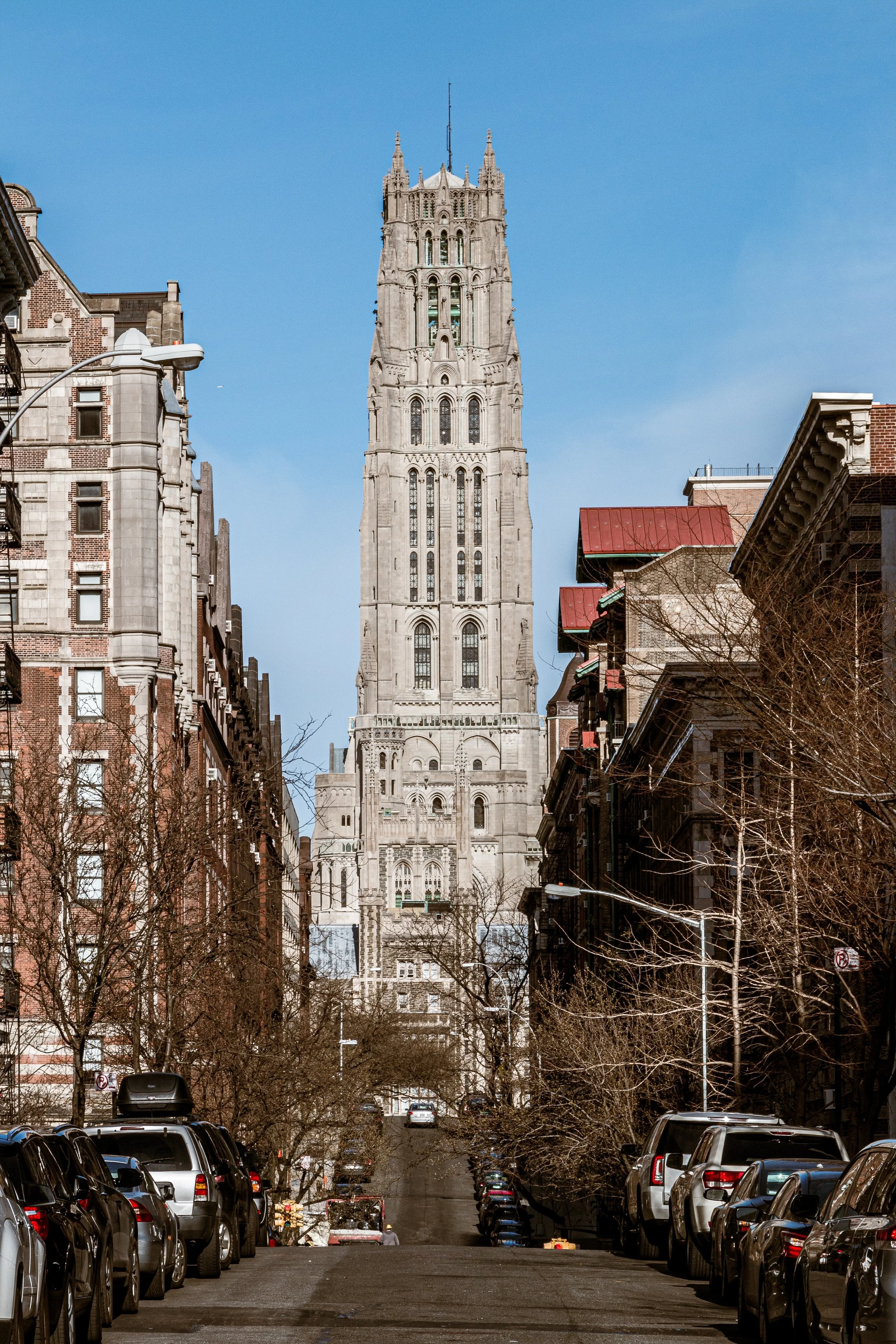 A city street with parked cars and leafless trees, leading to a prominent tall stone clock tower against a blue sky.