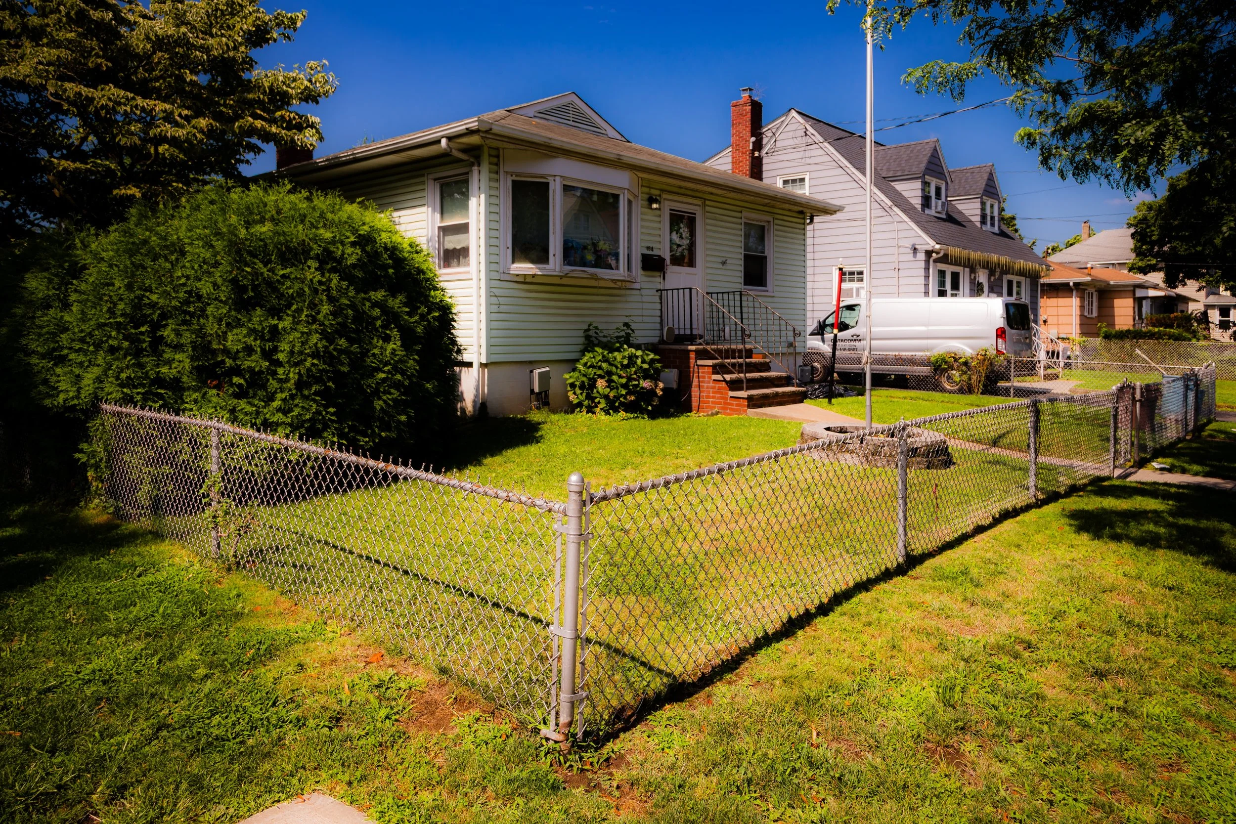 A small, light-colored house with a front porch, steps leading up to the door, and a lawn enclosed by a metal chain-link fence. There are bushes and trees around the house, and a parked white van nearby under a clear blue sky.