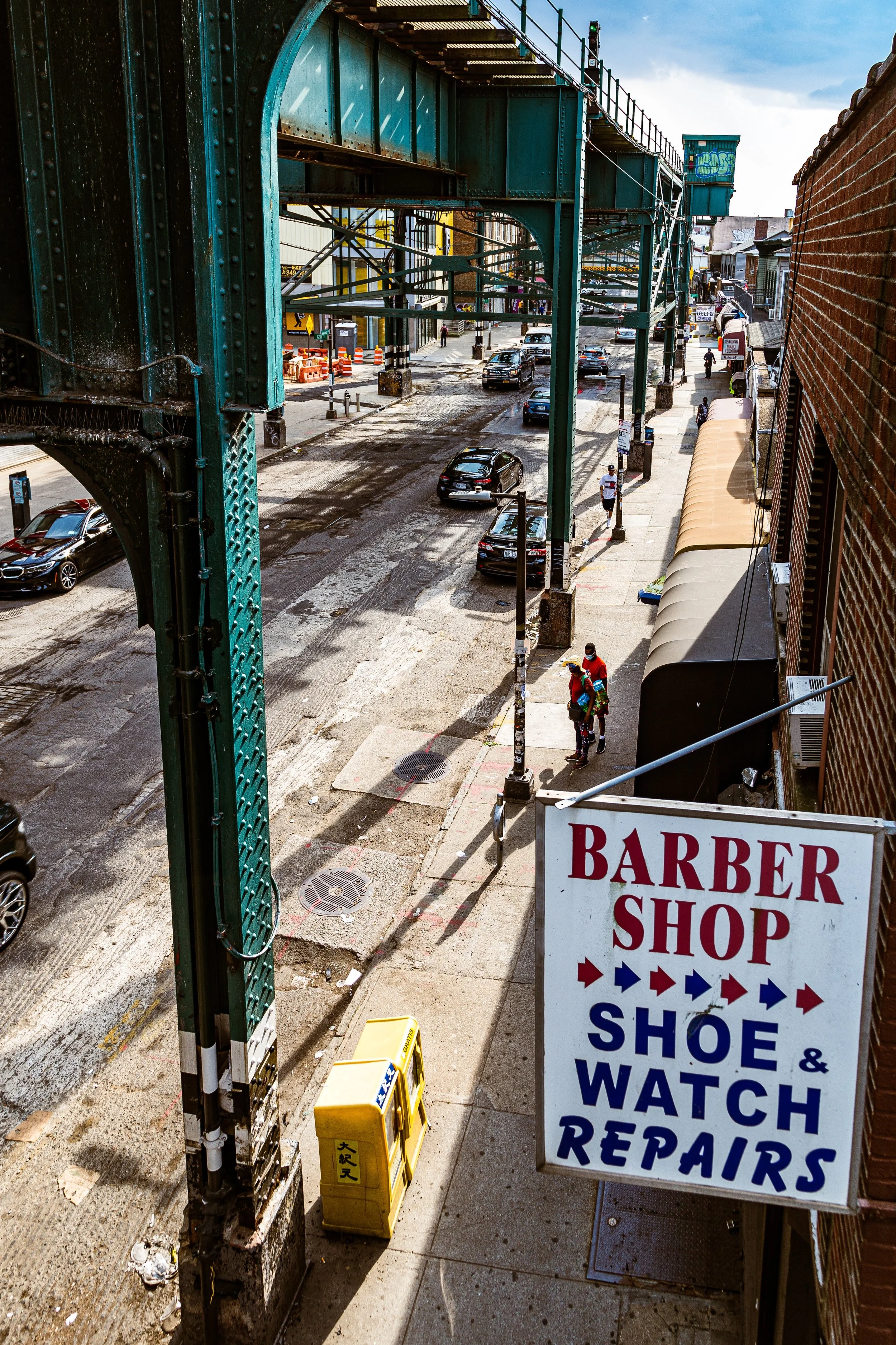 Street view showing cars on the road, pedestrians, an elevated train track, and storefronts including a sign for a barber shop that also offers shoe and watch repairs.