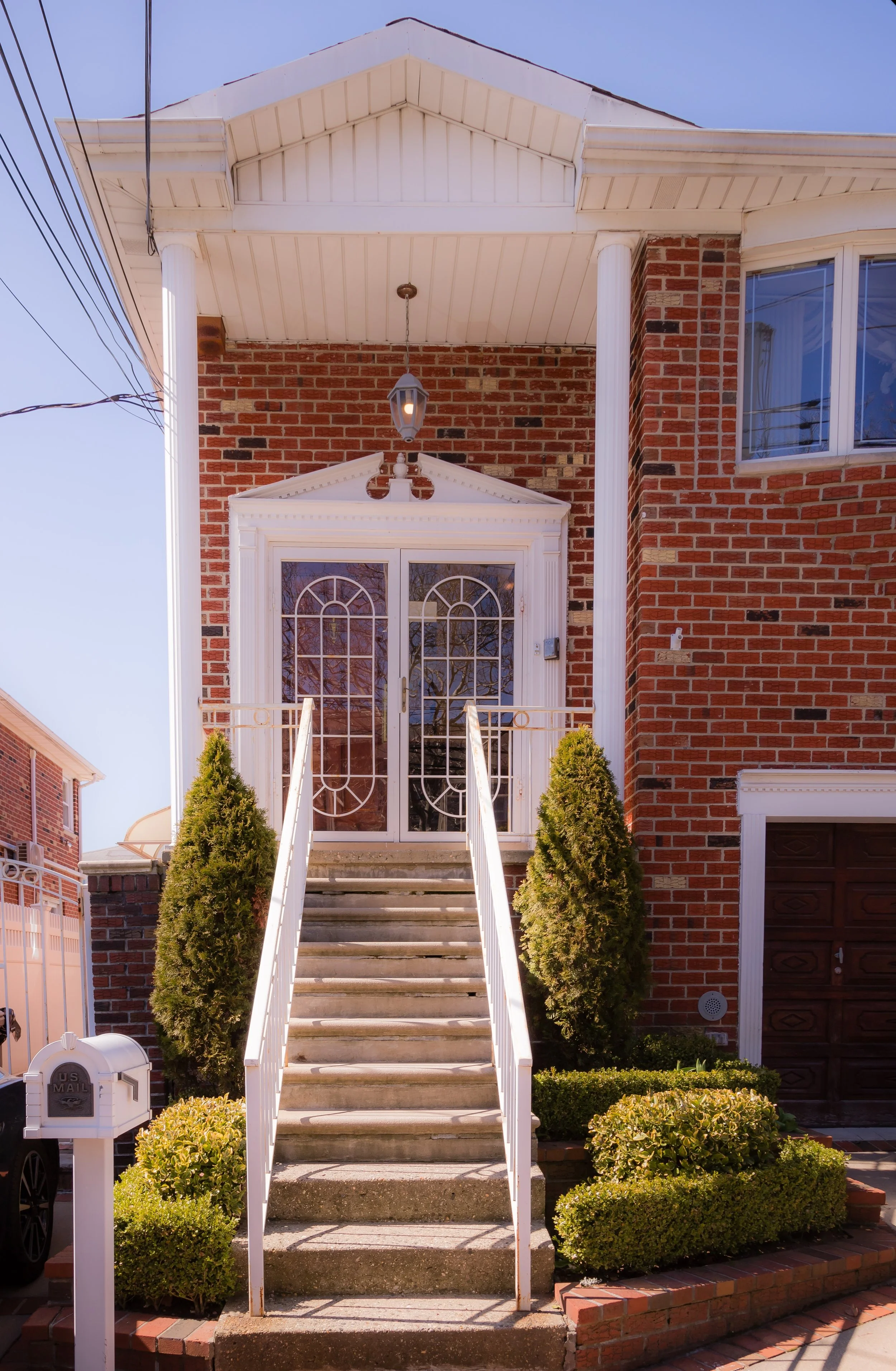 Front view of a two-story brick house with stairs leading to a front door with decorative glass panels. The house has white trim and two green shrubs flanking the stairs, with a mailbox in the front yard.