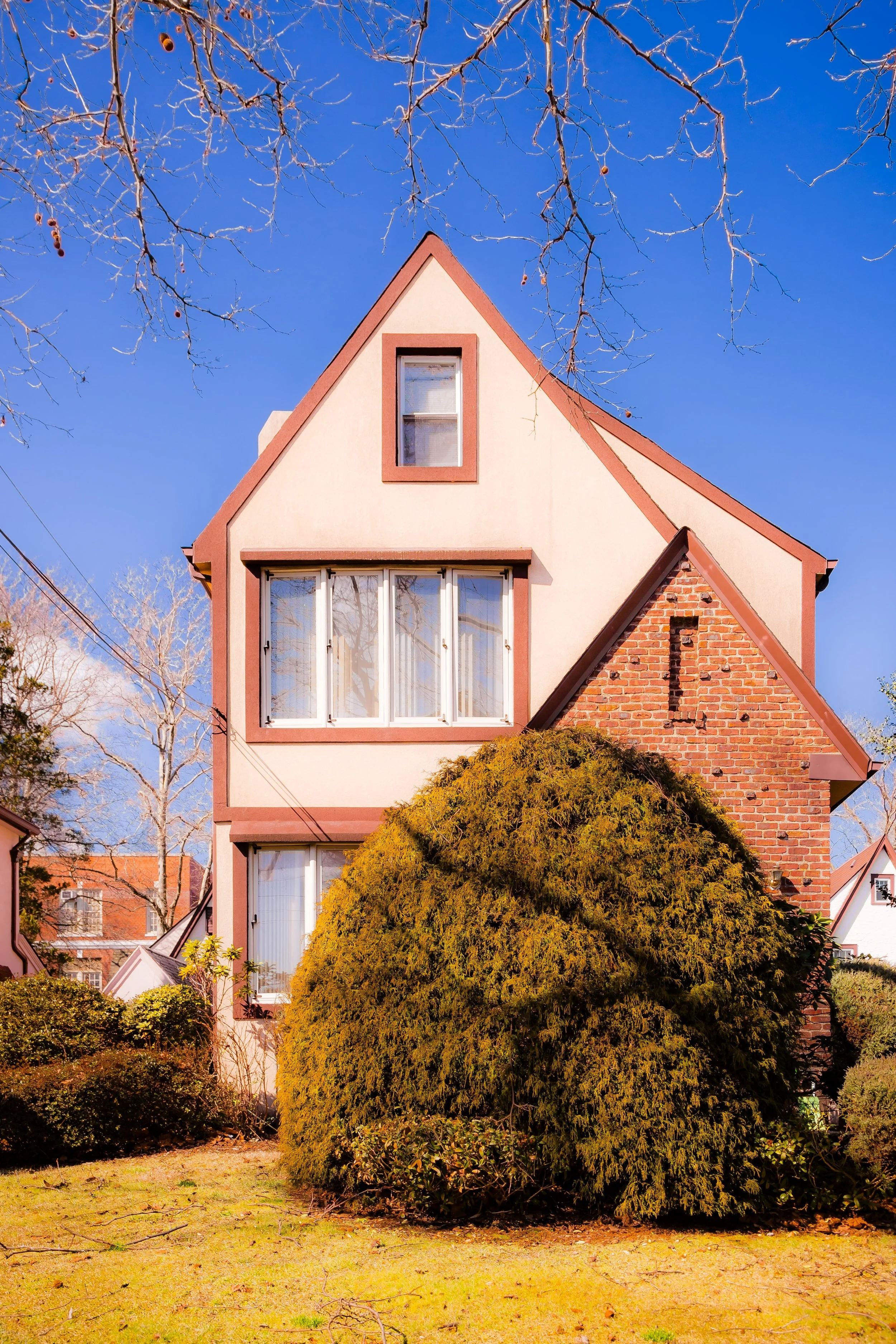 A two-story house with a steep roof and large front windows, surrounded by trees and bushes, under a clear blue sky.