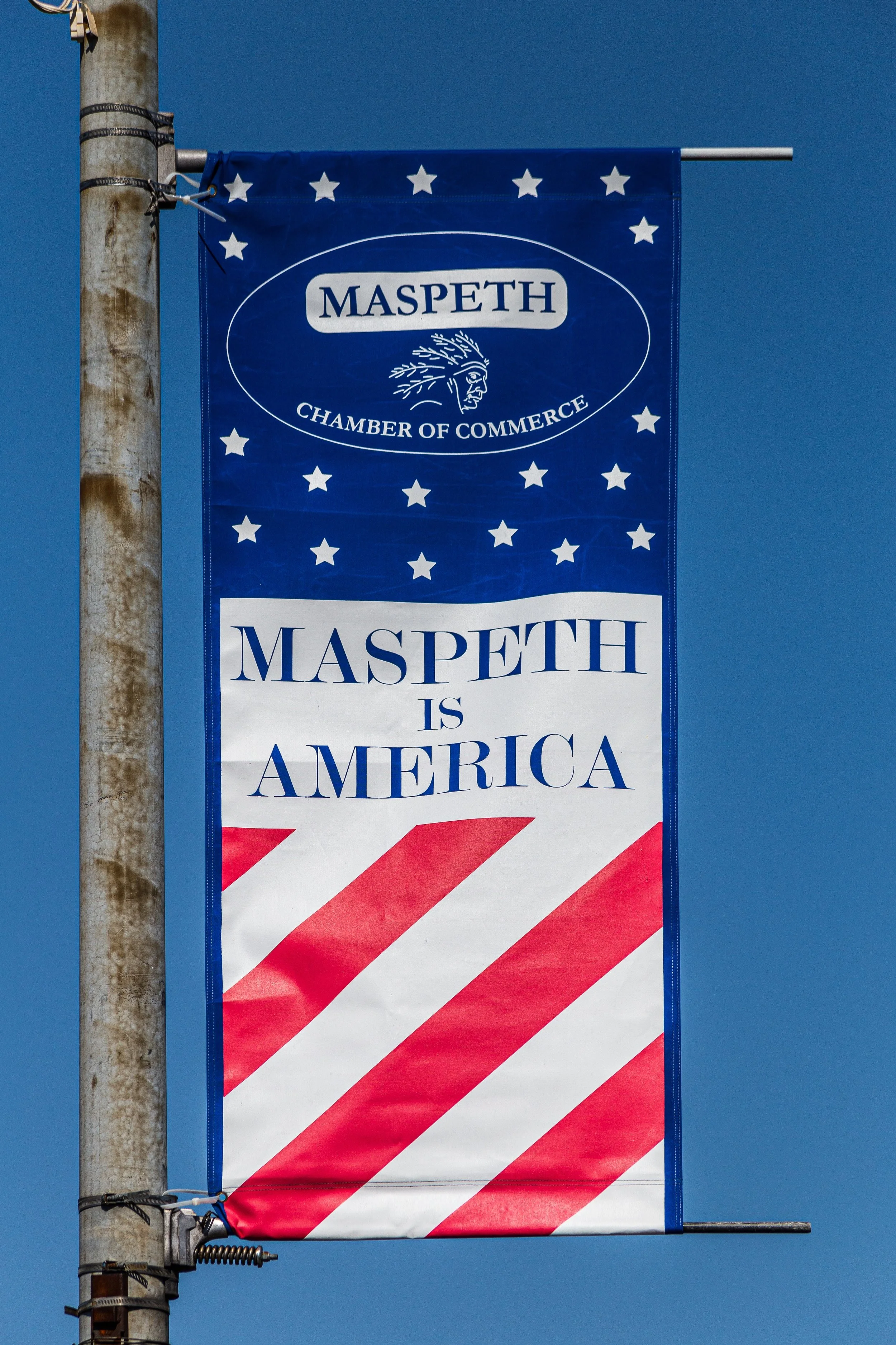 A patriotic banner on a pole that reads "MASPETH IS AMERICA" with a design of red and white stripes at the bottom, stars, and the Maspeth Chamber of Commerce logo at the top.