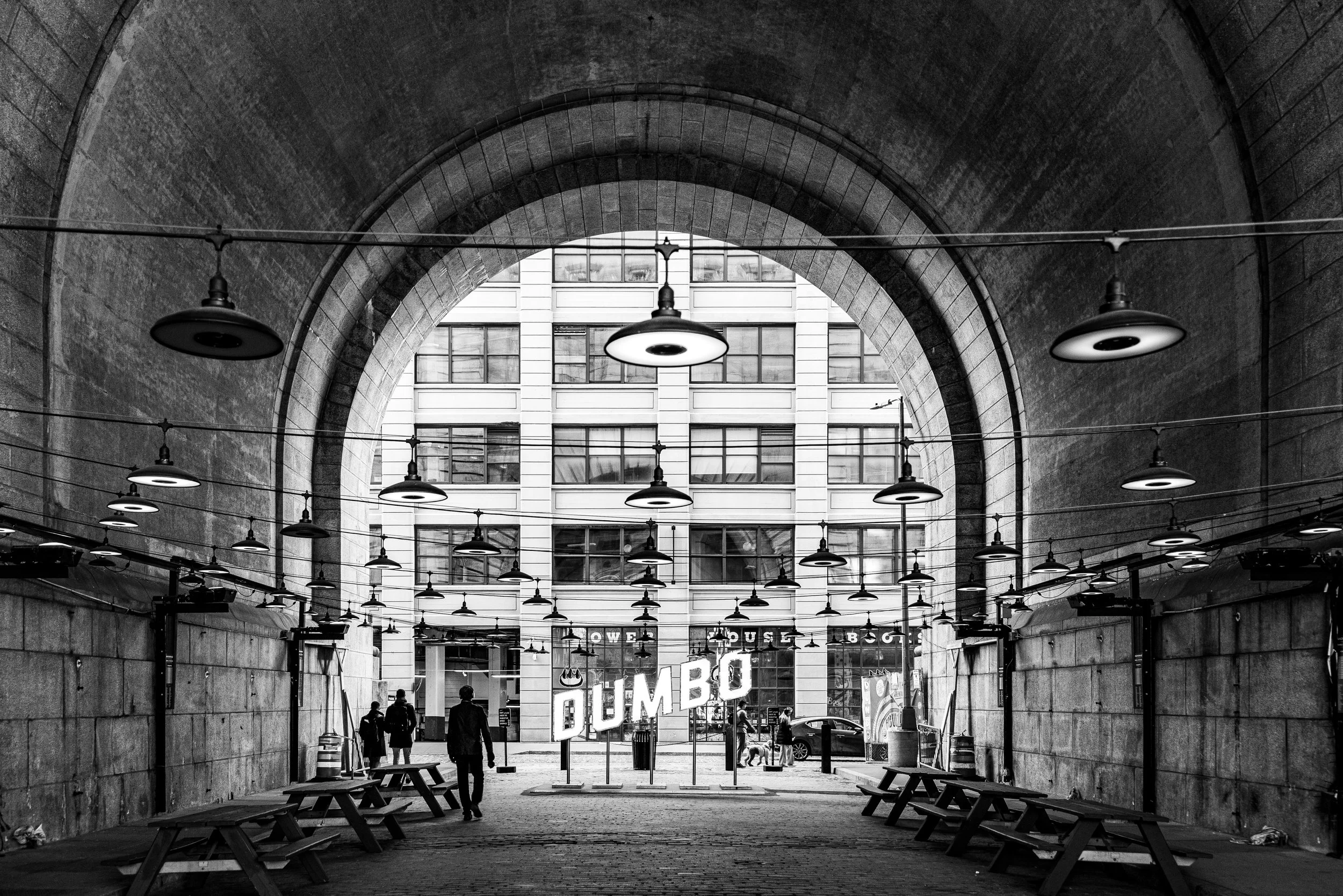 Black and white photograph of an urban underpass with a view towards a modern building and a neon "DUMBO" sign on the street level.