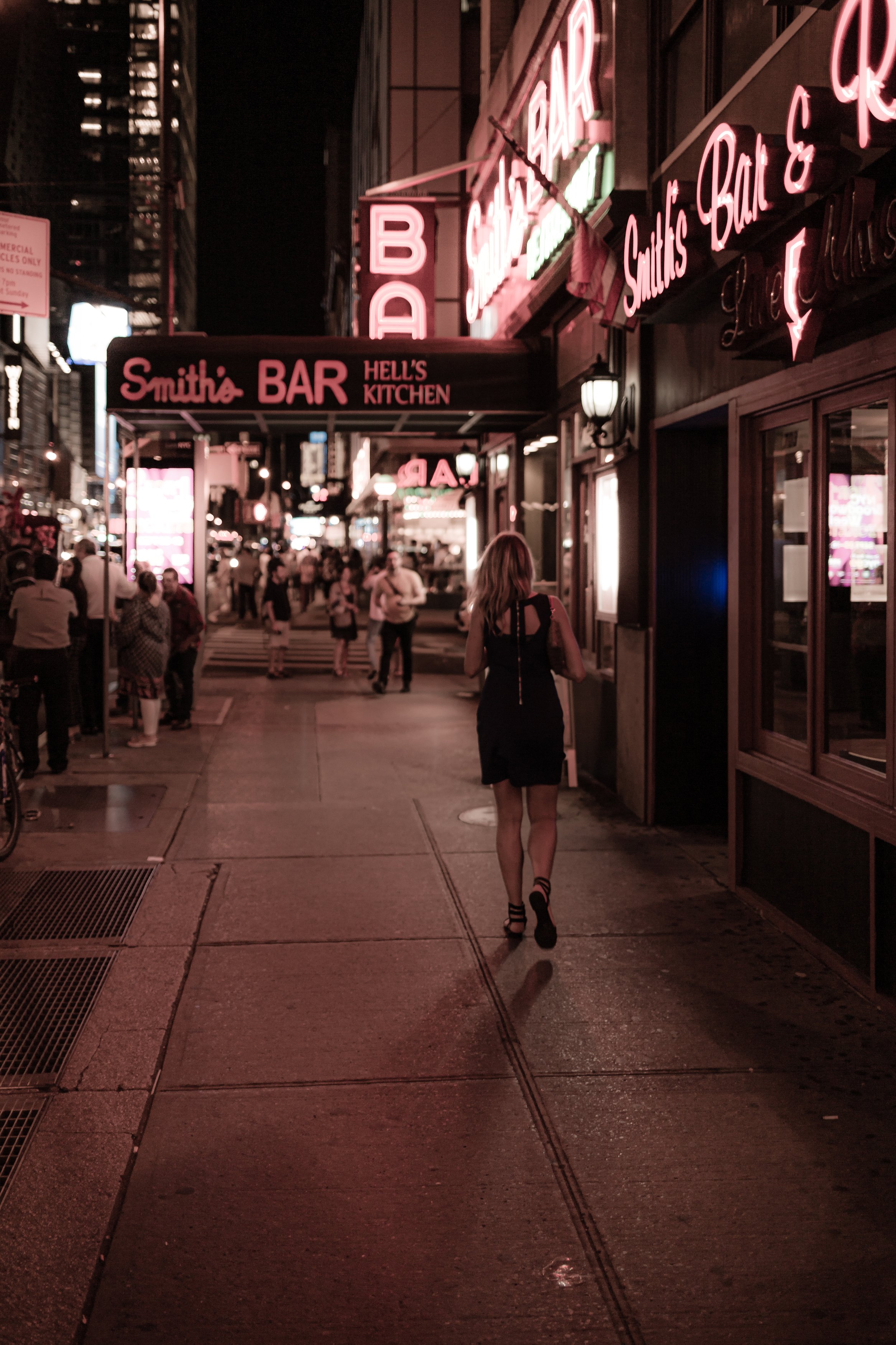 A woman walks alone on a city sidewalk at night, illuminated by pink neon signs that read 'Smith's BAR' and 'HELL'S KITCHEN.' Several people are visible further down the street, and the scene has an urban nightlife atmosphere.