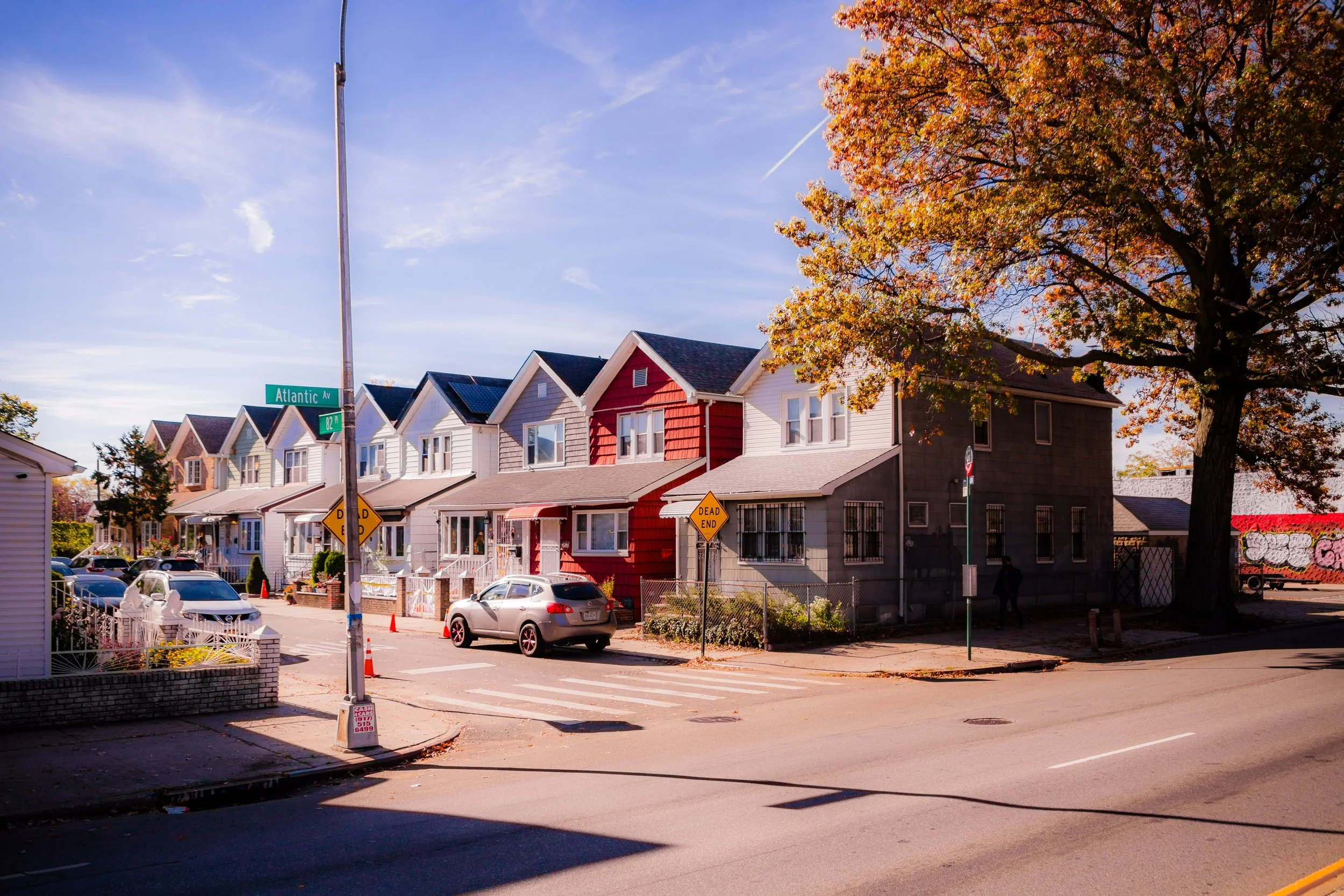 A street scene in a neighborhood with a row of colorful houses, cars parked along the street, and a large tree with fall foliage on the right side. The street sign indicates Atlantic Avenue and 82nd. There are traffic cones and a 'Dead End' sign visible.