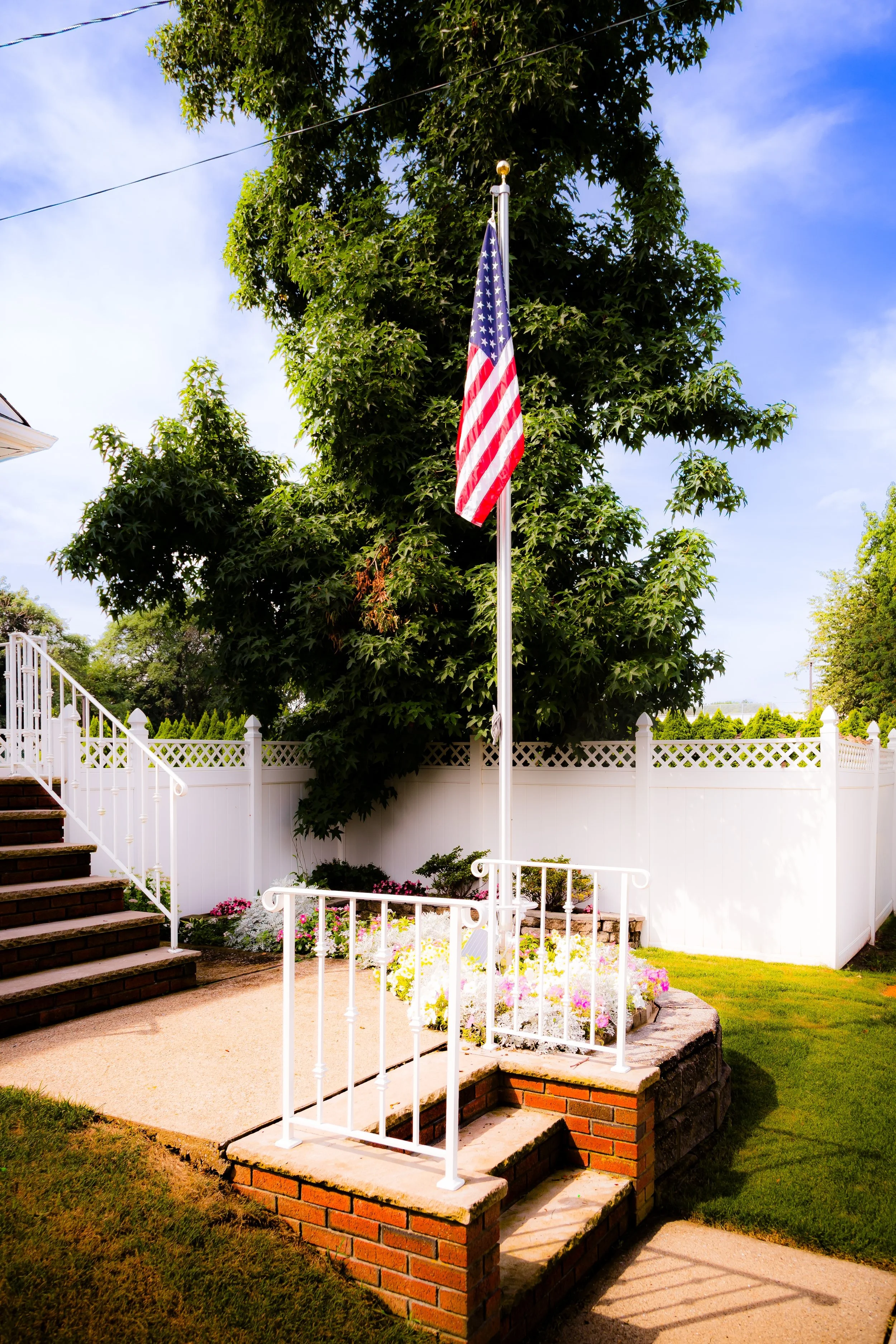 American flag on a flagpole in a backyard with a white fence, green trees, and a flower bed.