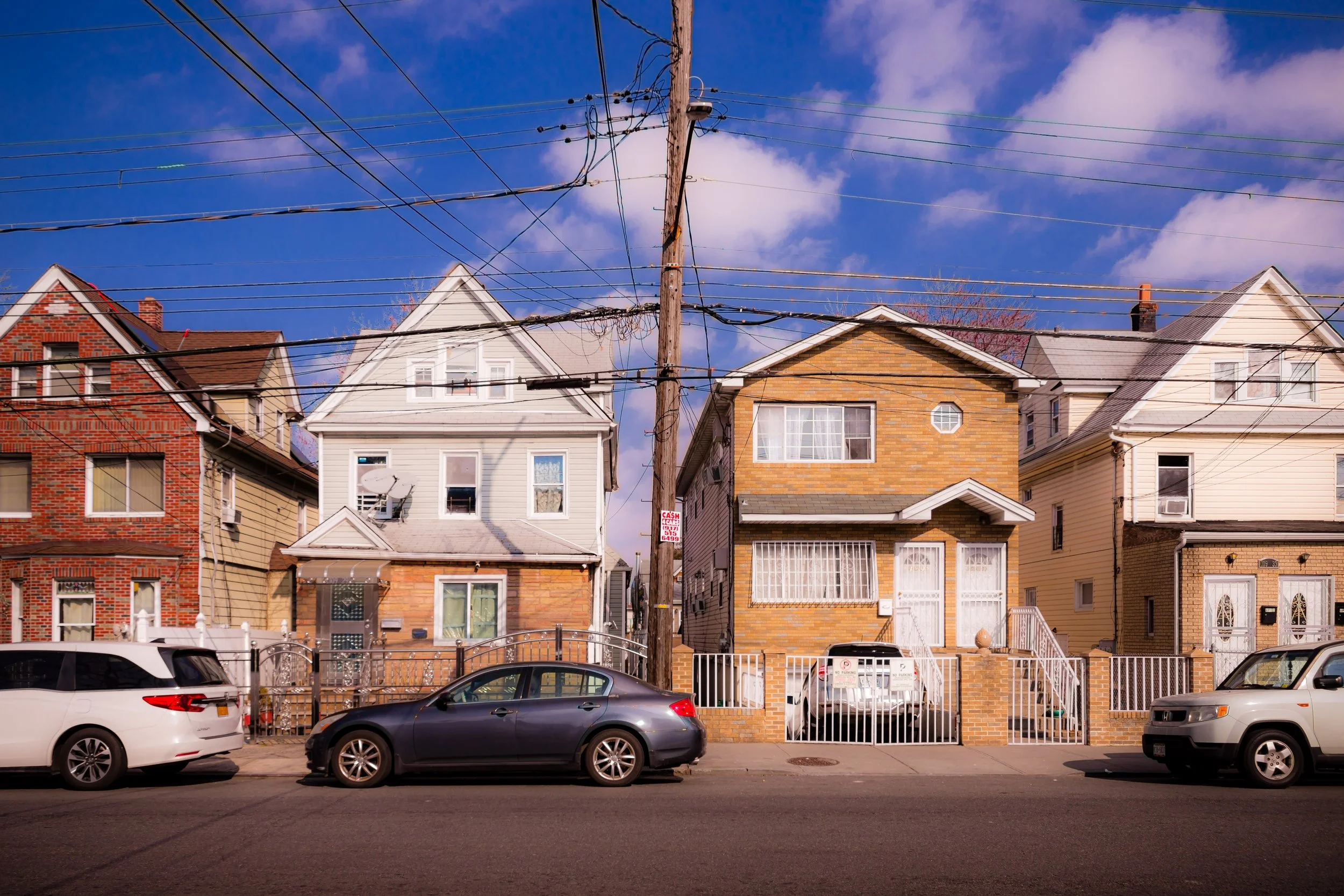 A row of houses on a city street with parked cars in front, utility pole and overhead wires, and a blue sky with scattered clouds.