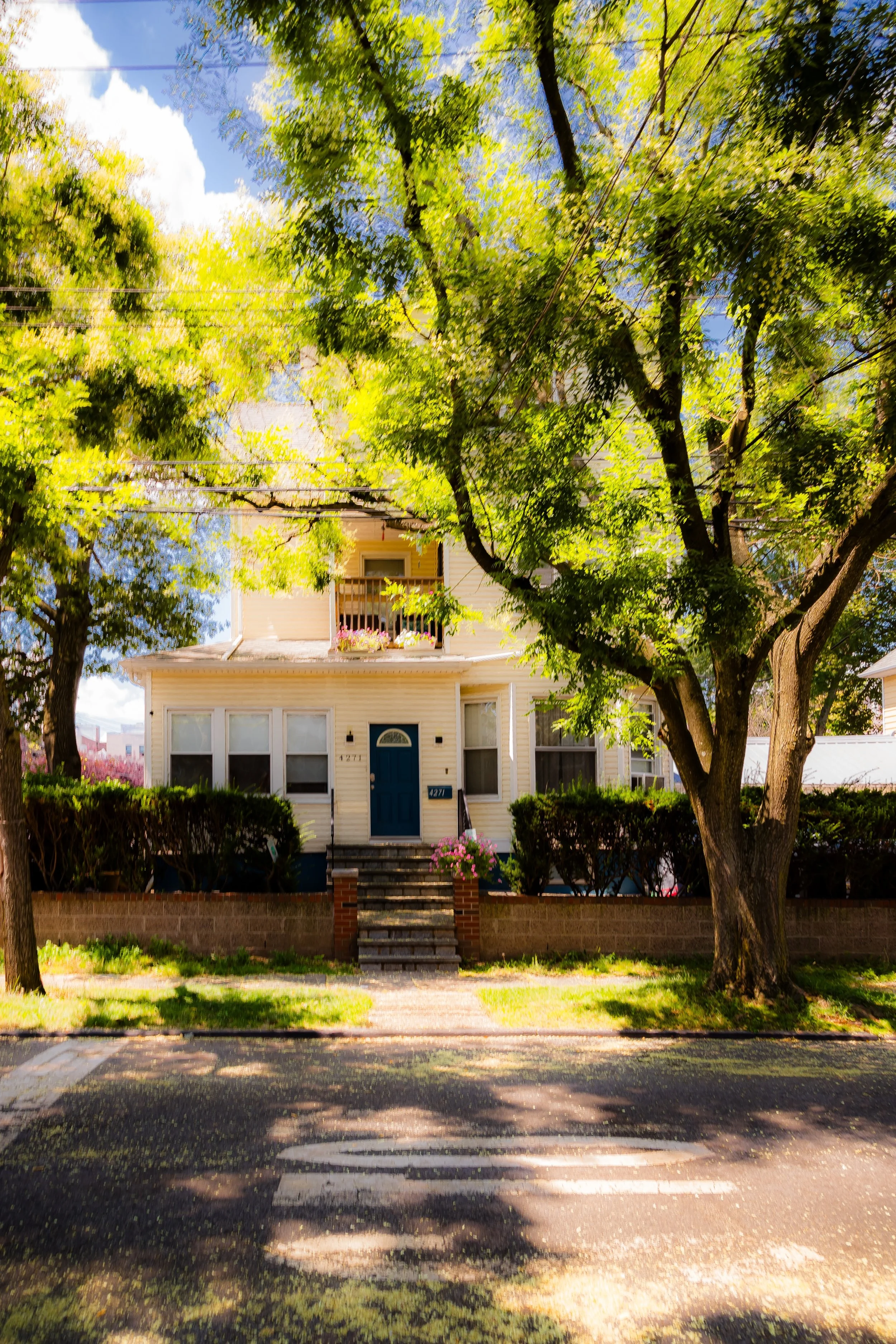 A yellow house with a blue front door, surrounded by trees with green leaves, and a brick staircase leading to the entrance. A sidewalk and crosswalk are in the foreground.