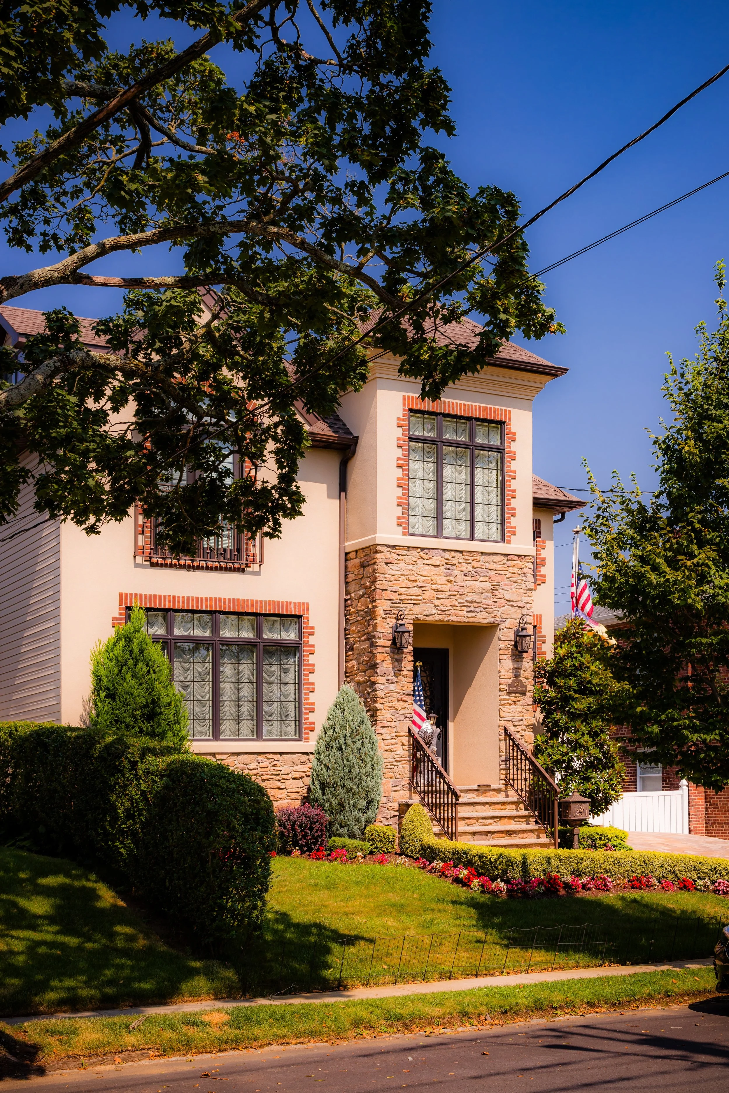 A two-story house with a stone and stucco exterior, large windows with curtains, and a front door with stairs. The house is surrounded by well-maintained green bushes and colorful flowers, with American flags near the entrance. There are trees and power lines in the foreground and a bright blue sky overhead.