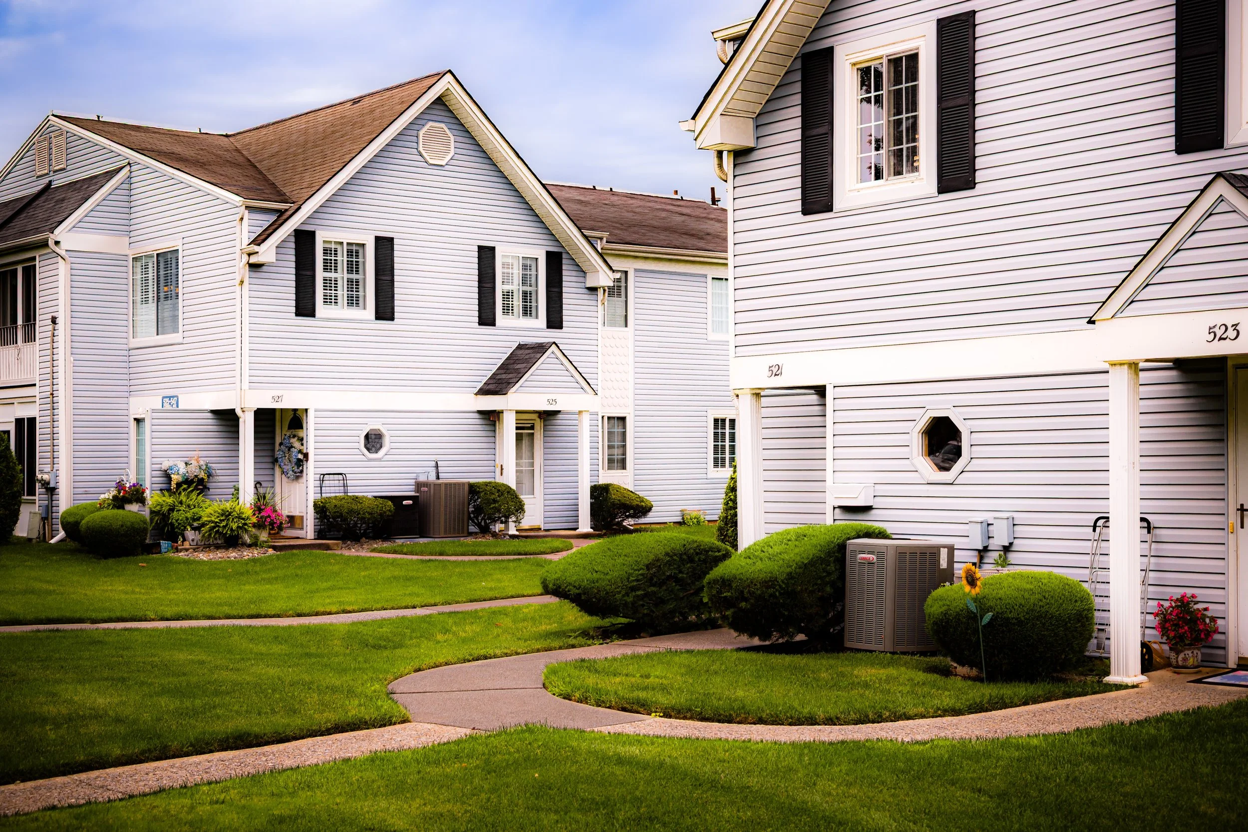 View of a row of white townhouses with black shutters, green lawns, and neatly trimmed bushes. There are small walkways, potted plants, and outdoor air conditioning units.