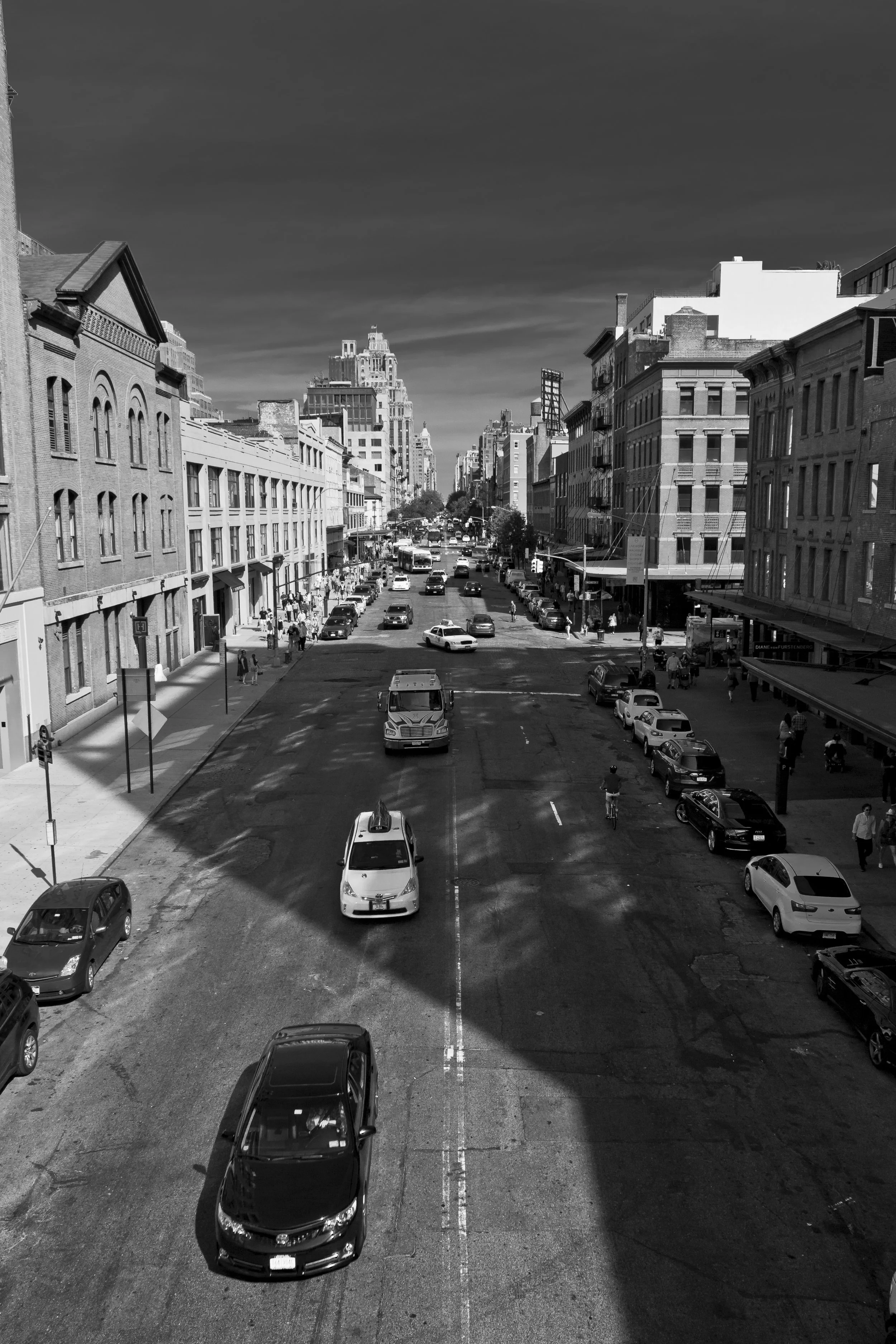 Black and white photo of a city street with parked cars and buildings on both sides, leading to taller buildings in the background under a partly cloudy sky.