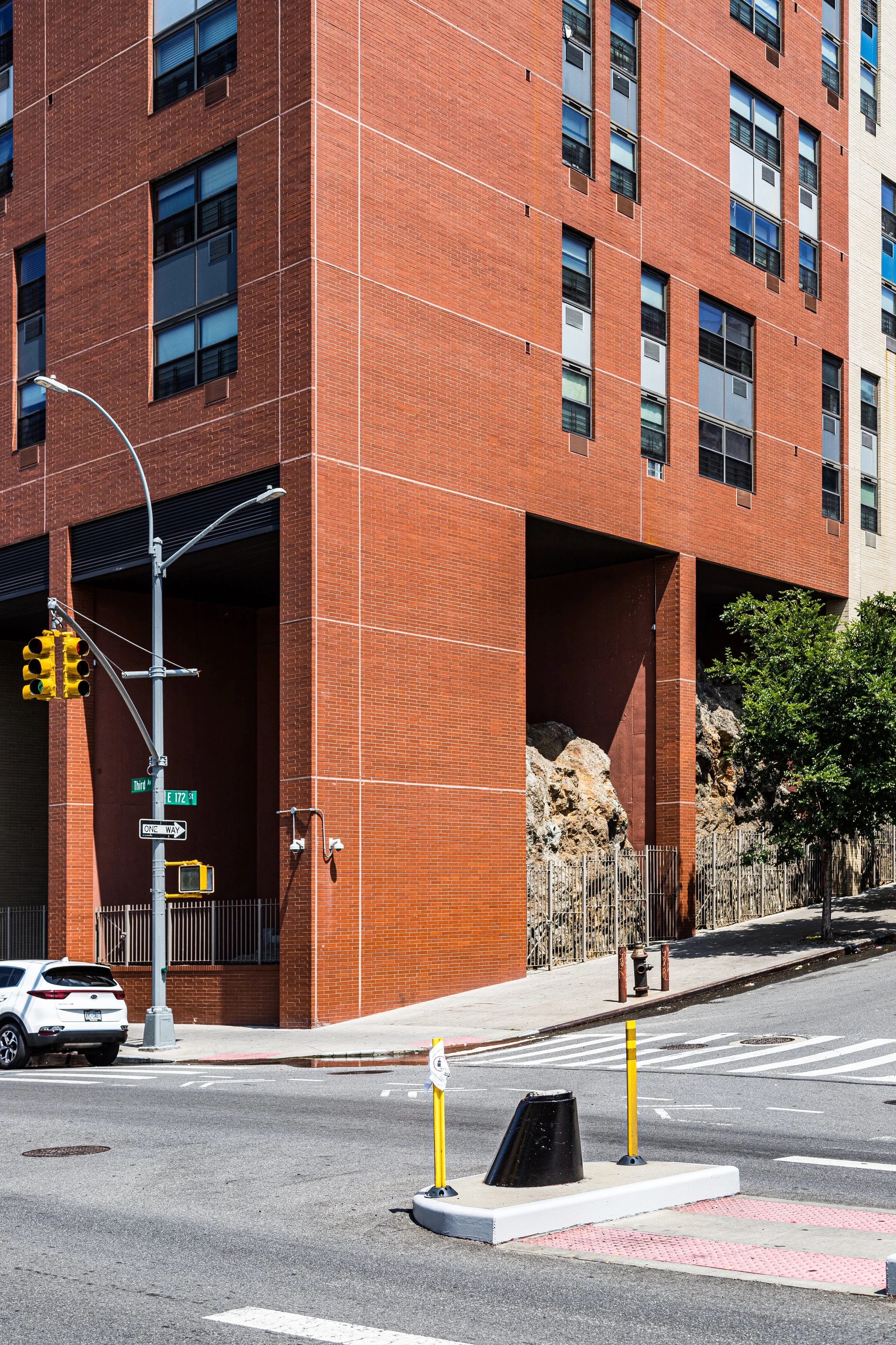 A modern red brick multi-story building with multiple windows, situated on a city street corner. There is a traffic light and street signs indicating 'Third Street' and 'E 172nd Street', along with a yellow pedestrian crossing button. A white SUV is parked nearby, and there are trees and rocks on the sidewalk.