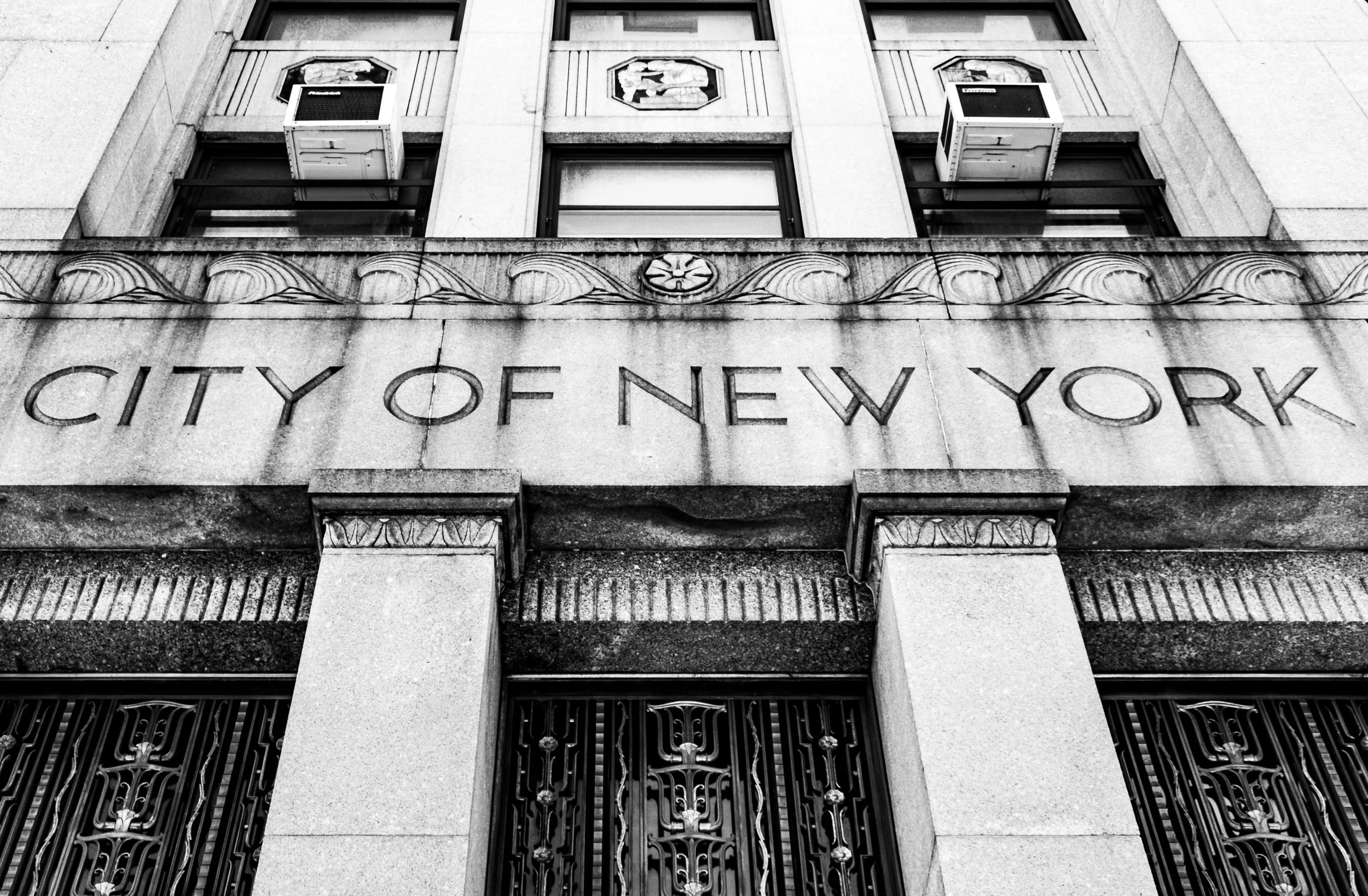 Black and white photograph of the front of a building with the words 'City of New York' engraved above the entrance. The building features decorative architectural elements and two windows with air conditioning units.
