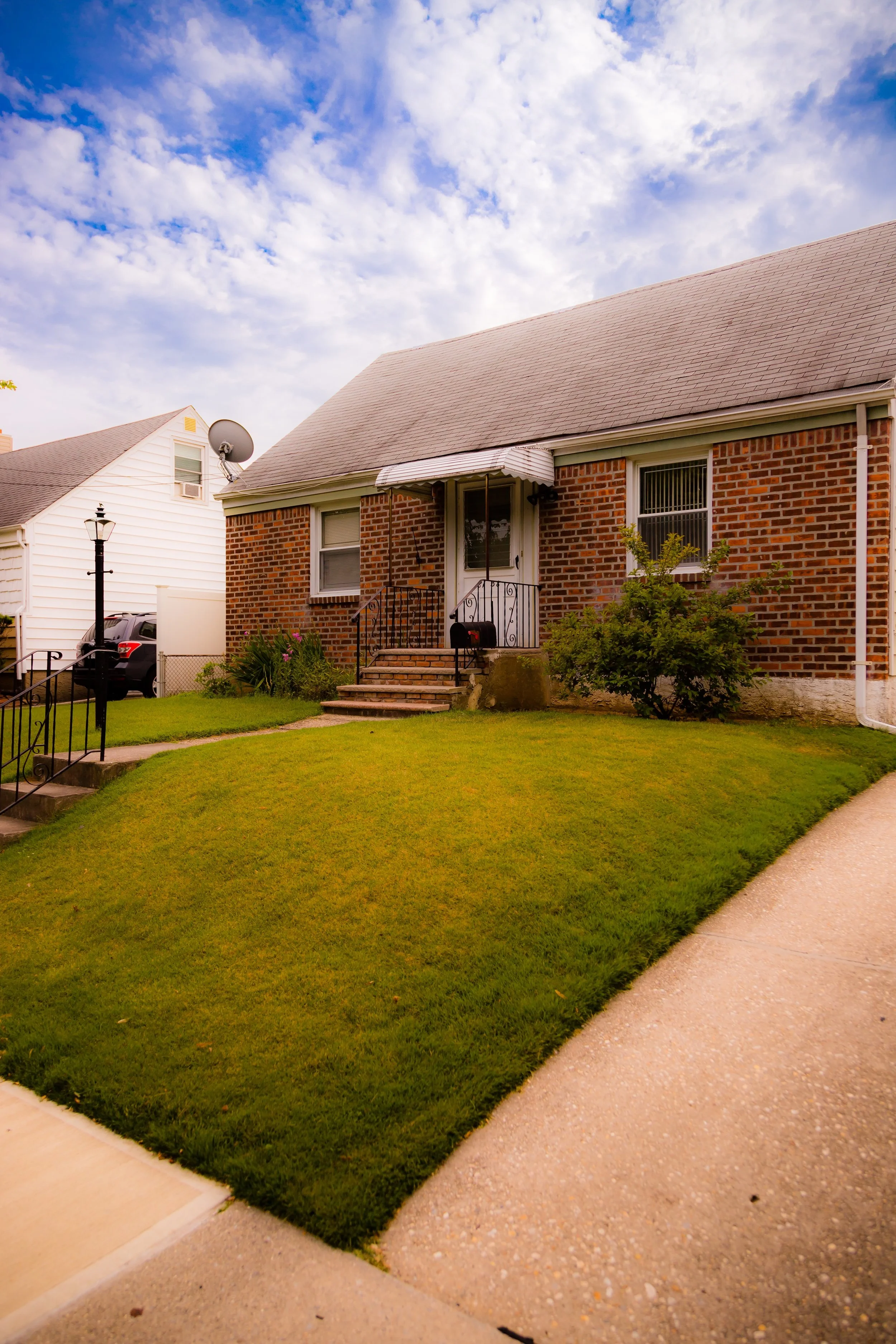 Front yard of a brick house with steps leading to the front door, surrounded by a green lawn and a sidewalk, under a partly cloudy sky.