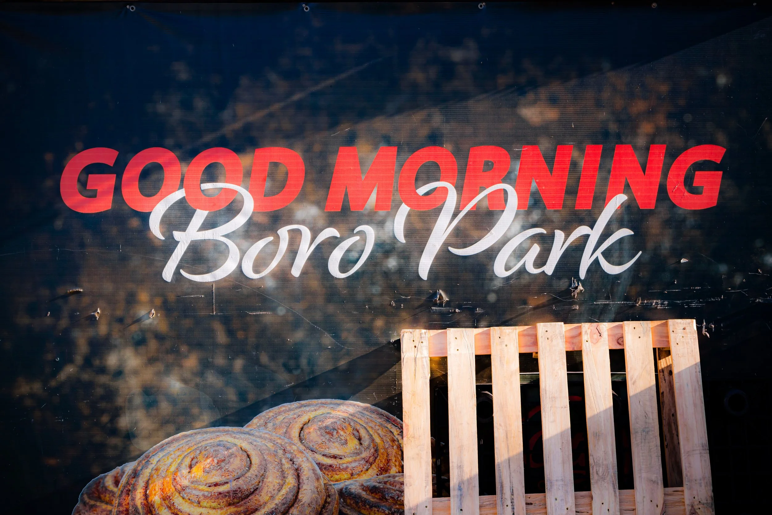 A sign on a glass window reads 'Good Morning Boro Park' with the background showing pastries and a wooden pallet.