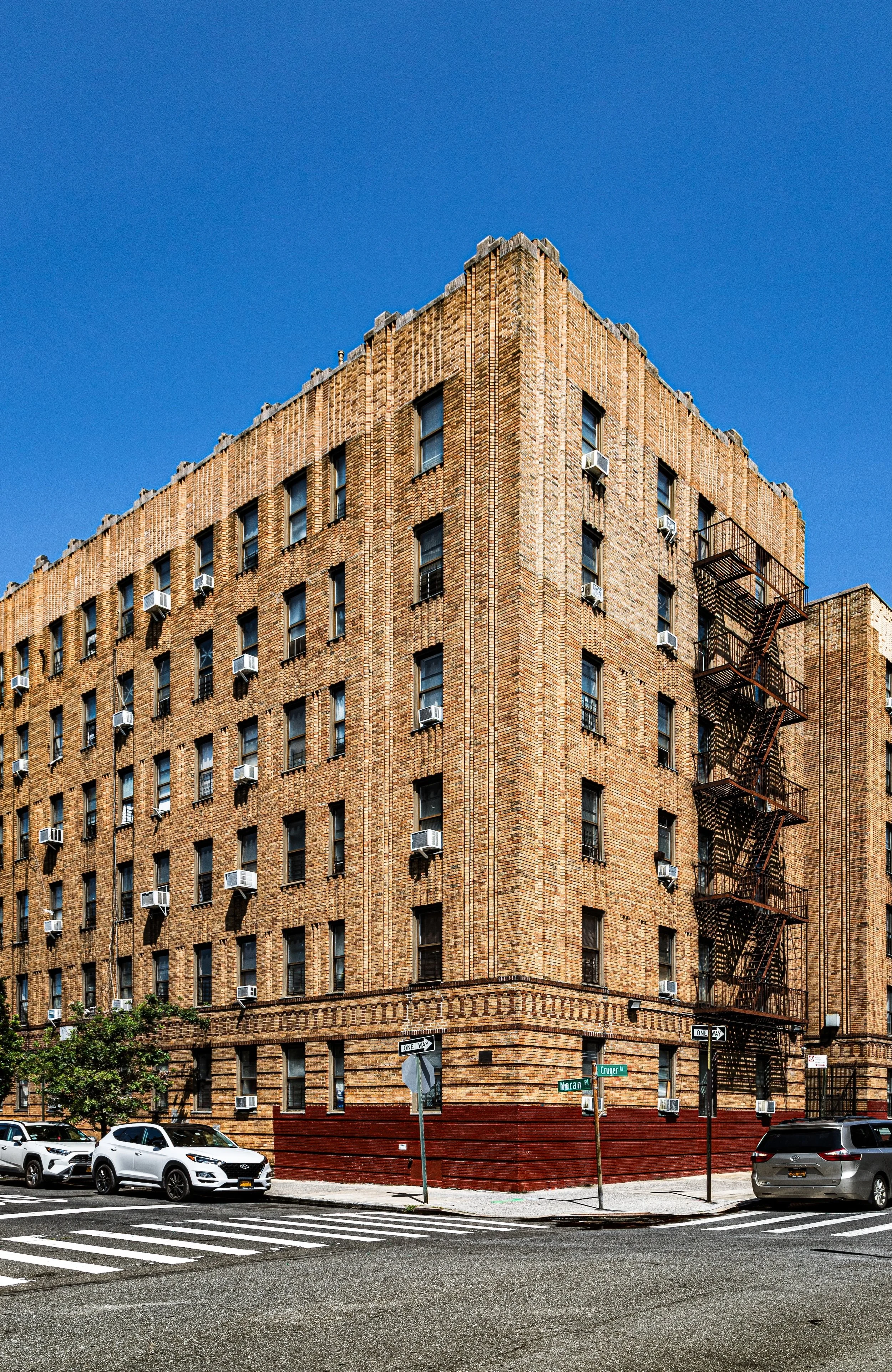 A multi-story brick building on a city street corner with several windows, air conditioning units, and a black fire escape on the side. Cars are parked in front and street signs are visible at the intersection.