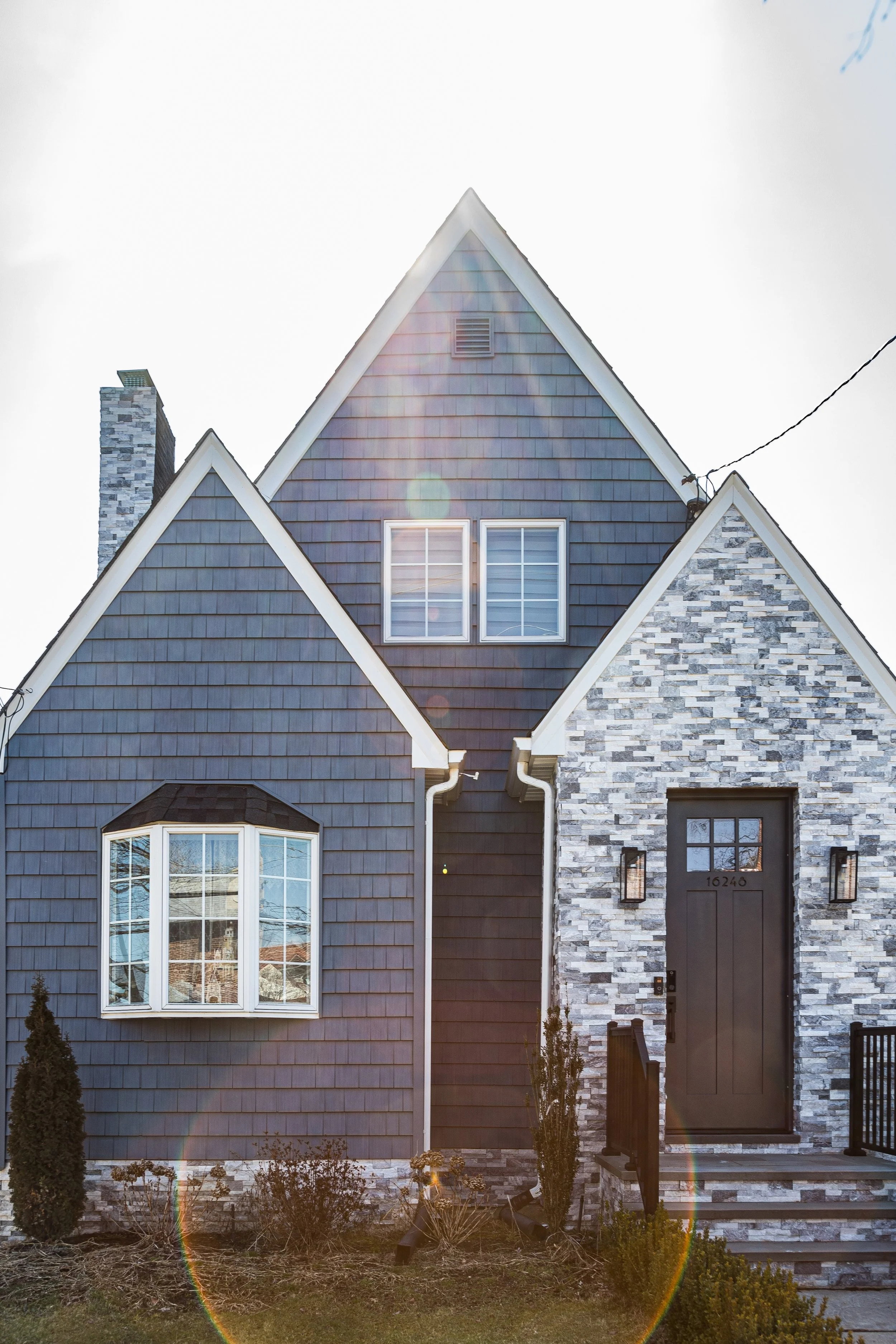 Front exterior of a modern house with gray siding, stone accents, a gabled roof, and a black front door with the number 16246.
