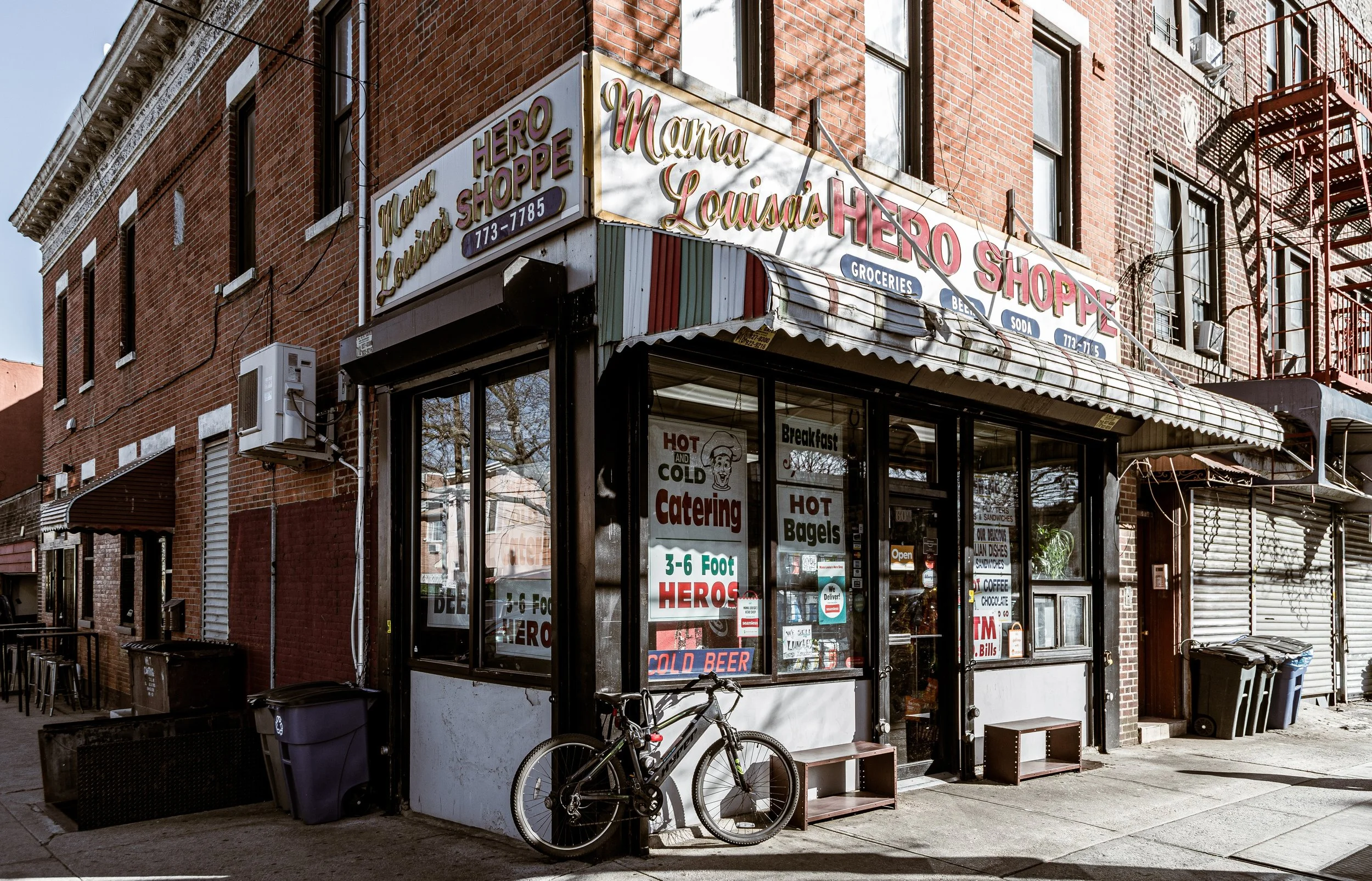 Street view of Mama Louisa's Hero Shoppe, a small grocery store in a brick building with signs for groceries, beer, soda, and catering, a bicycle parked outside, and an awning over the entrance.