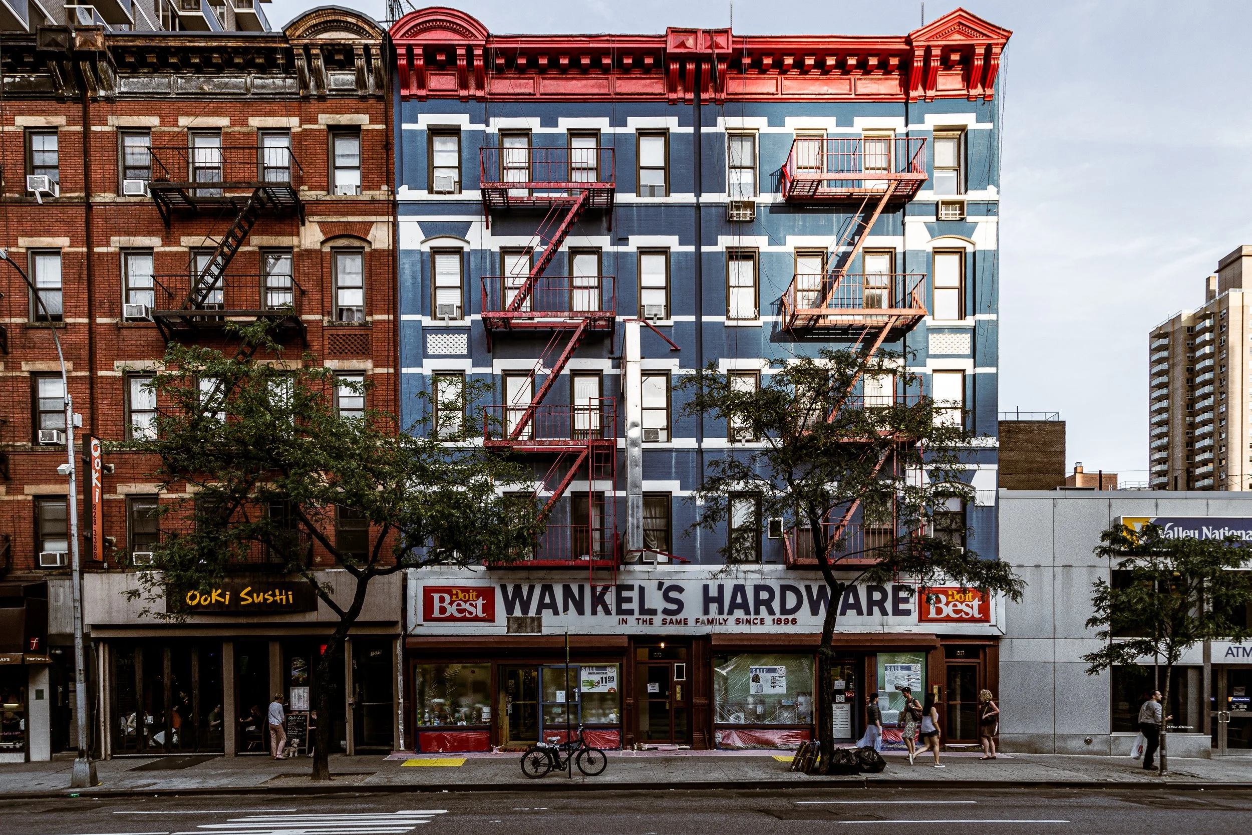 A colorful city building with red, blue, and white exterior paint and red fire escapes. The ground level has a hardware store called Wankel's Hardware, a sushi restaurant named Ooki Sushi, and an ATM. People walk on the sidewalk, and there are trees and parked bicycles in front.