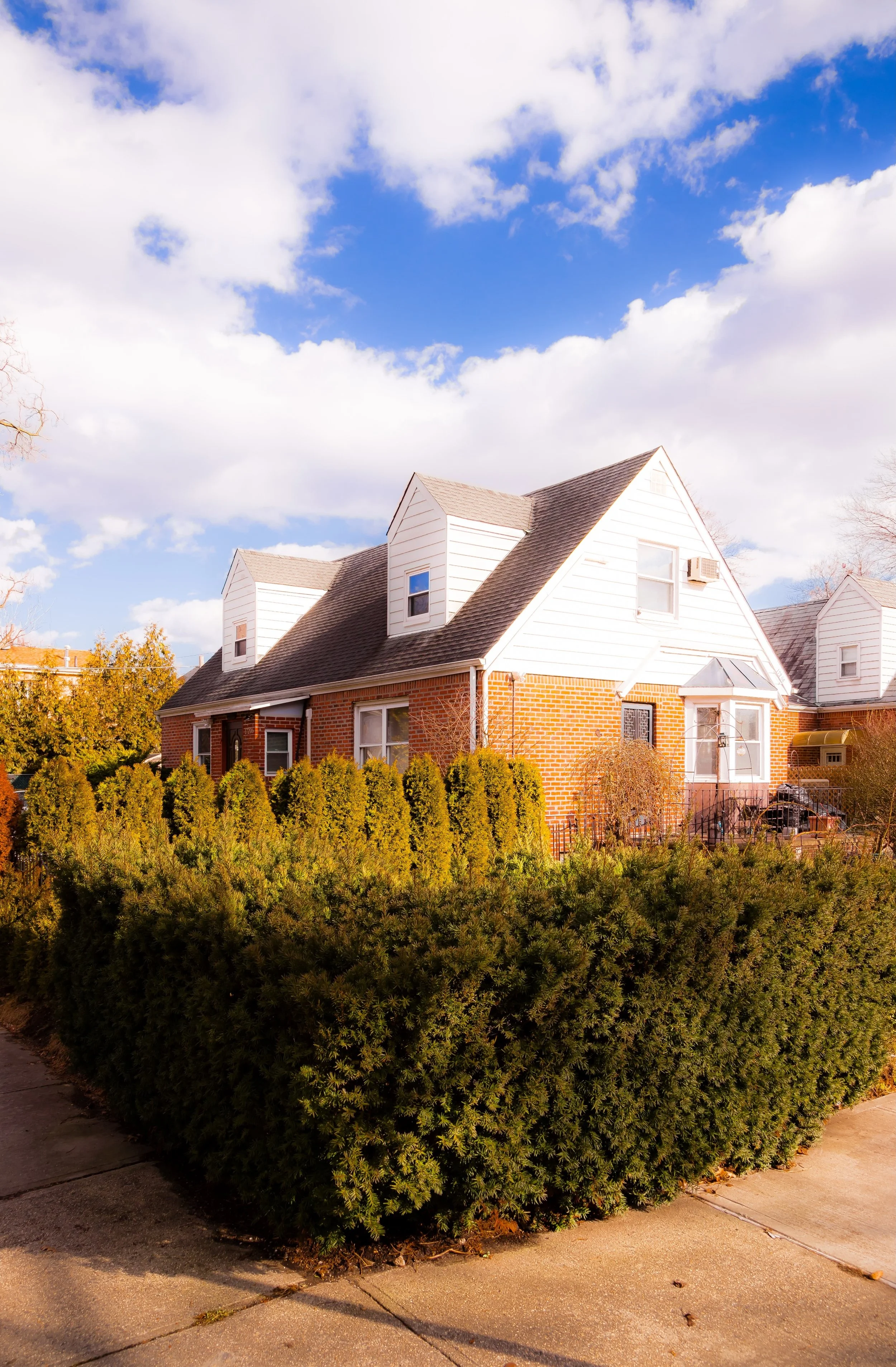 A suburban house with brick and white siding exterior, surrounded by well-manicured bushes, under a partly cloudy sky.