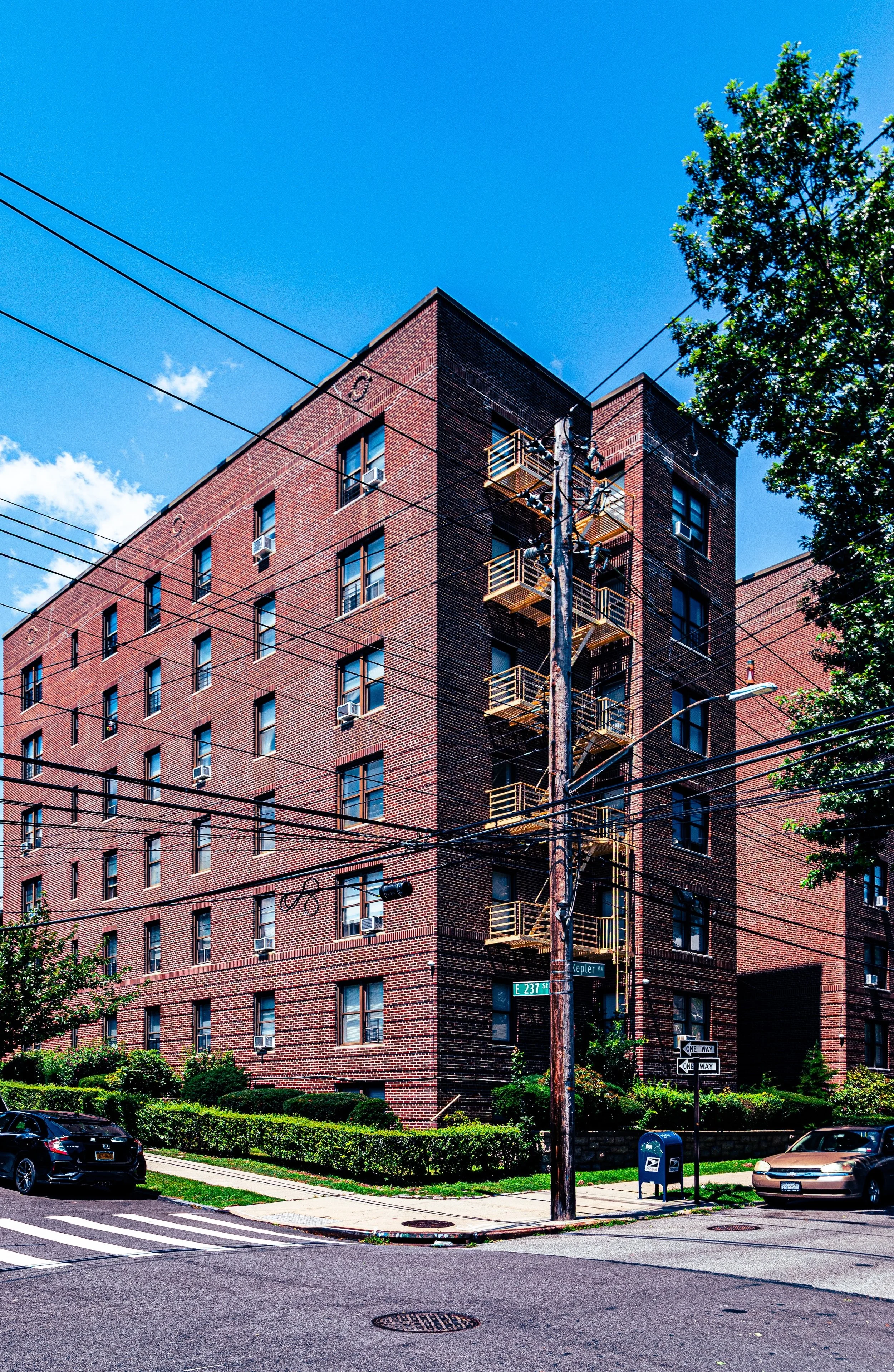 A multi-story red brick apartment building under a clear blue sky, with fire escape balconies on one side, power lines crossing in front, and cars parked along the street.