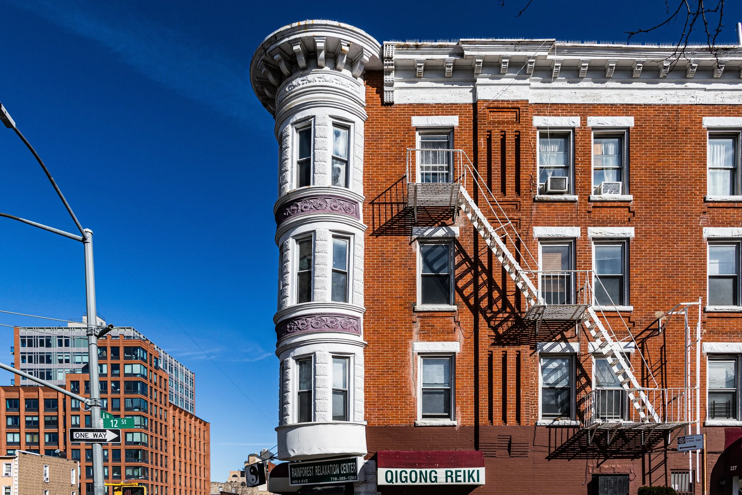 A red brick building with fire escape stairs on the front, adjacent to a rounded corner with white and purple decorative trim, and large windows. In the background is another modern building with many windows. There is blue sky overhead and street signs indicating 12th Street and one way.