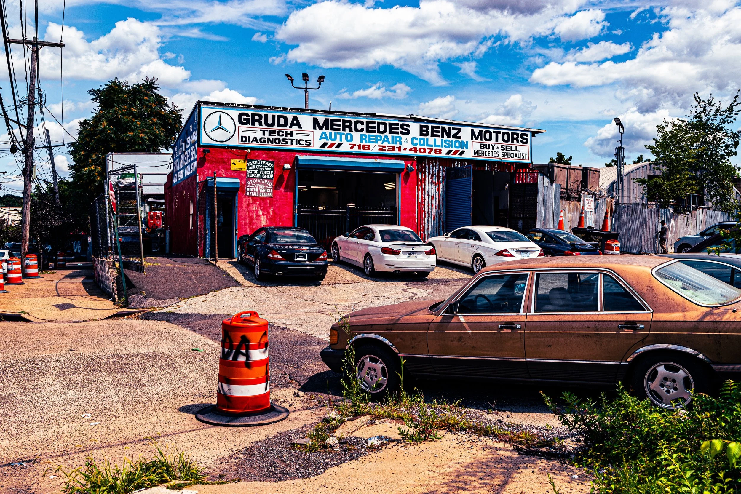A car repair shop with a sign that reads Gruda Mercedes Benz Motors, offering auto repair, collision, and diagnostics services. Several cars are parked outside, including a rusty vintage car in the foreground, under a partly cloudy sky.