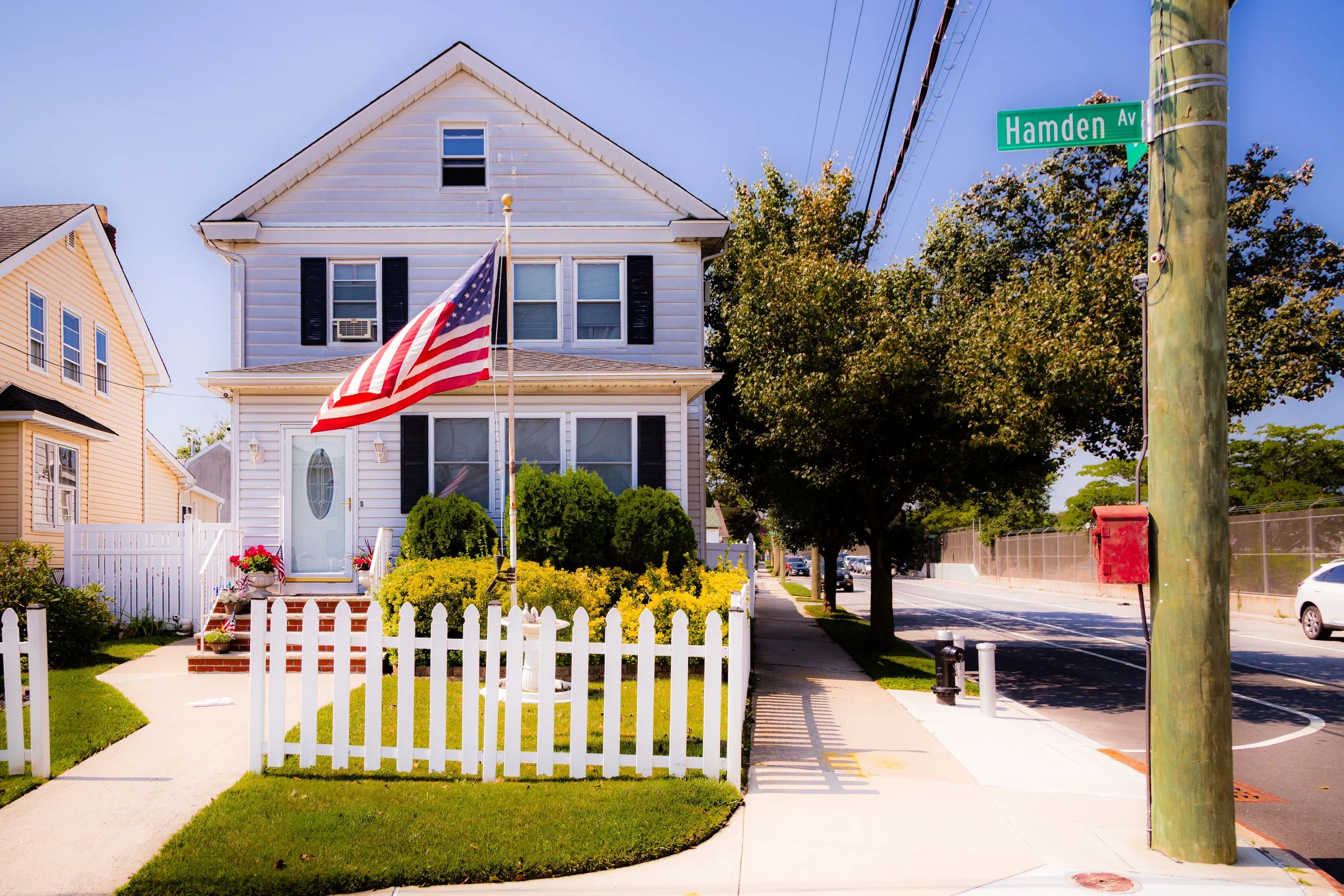 A white two-story house with a picket fence, American flag, and landscaped yard on a sunny street with trees and cars.