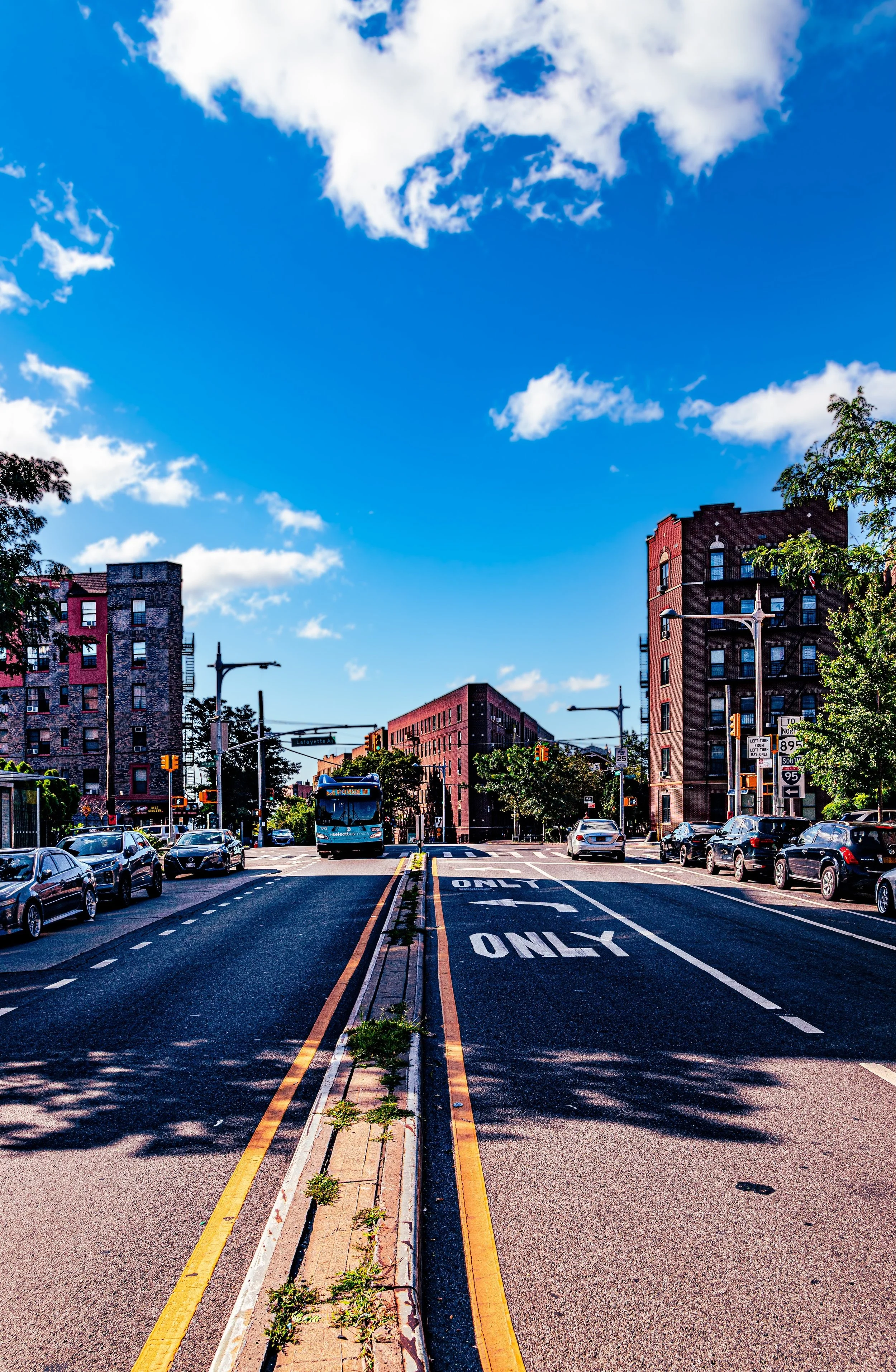 City street with cars parked on both sides, a bus in the middle, tall buildings under a blue sky with clouds, and road signs indicating directions.