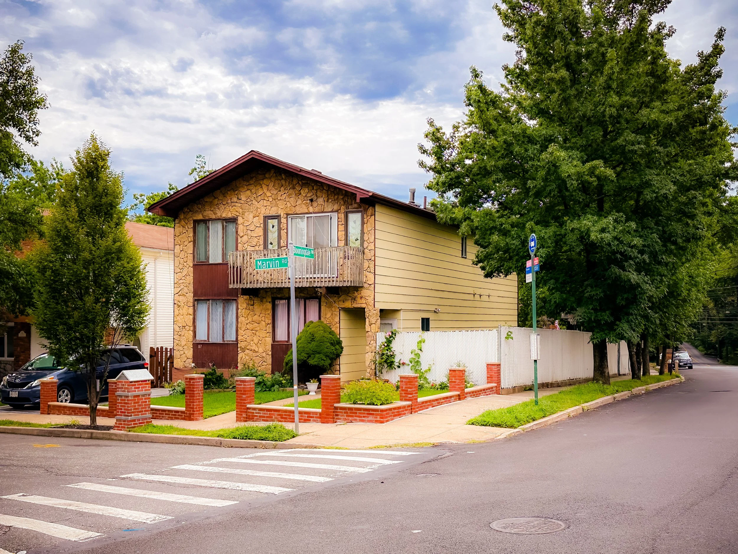 A two-story house at the corner of Marvin Road and Sherman Drive, featuring a stone facade on the front. There is a small front yard with bushes bordered by a low brick wall. The house has a balcony on the second floor and trees around the property. The street has a crosswalk and traffic signs visible.