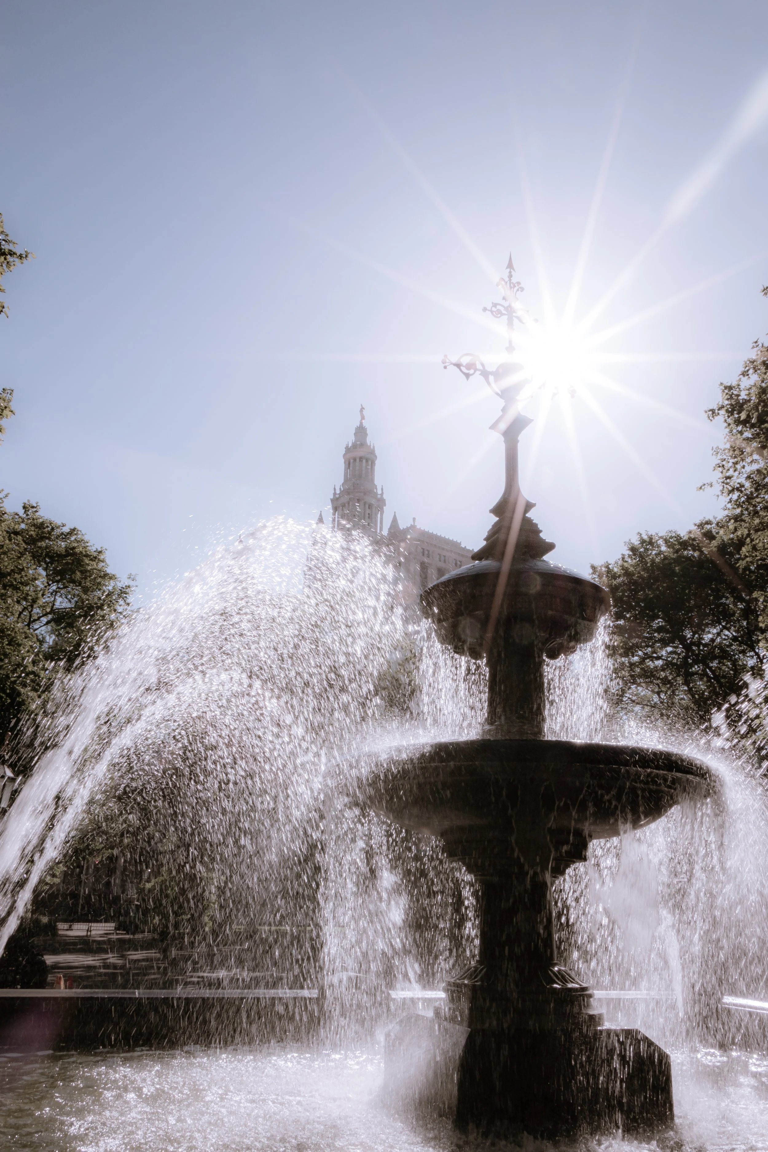A decorative fountain with water cascading down, sunlight shining brightly behind a tall ornate street lamp, and a historic building with a tower in the background on a clear day with blue sky.