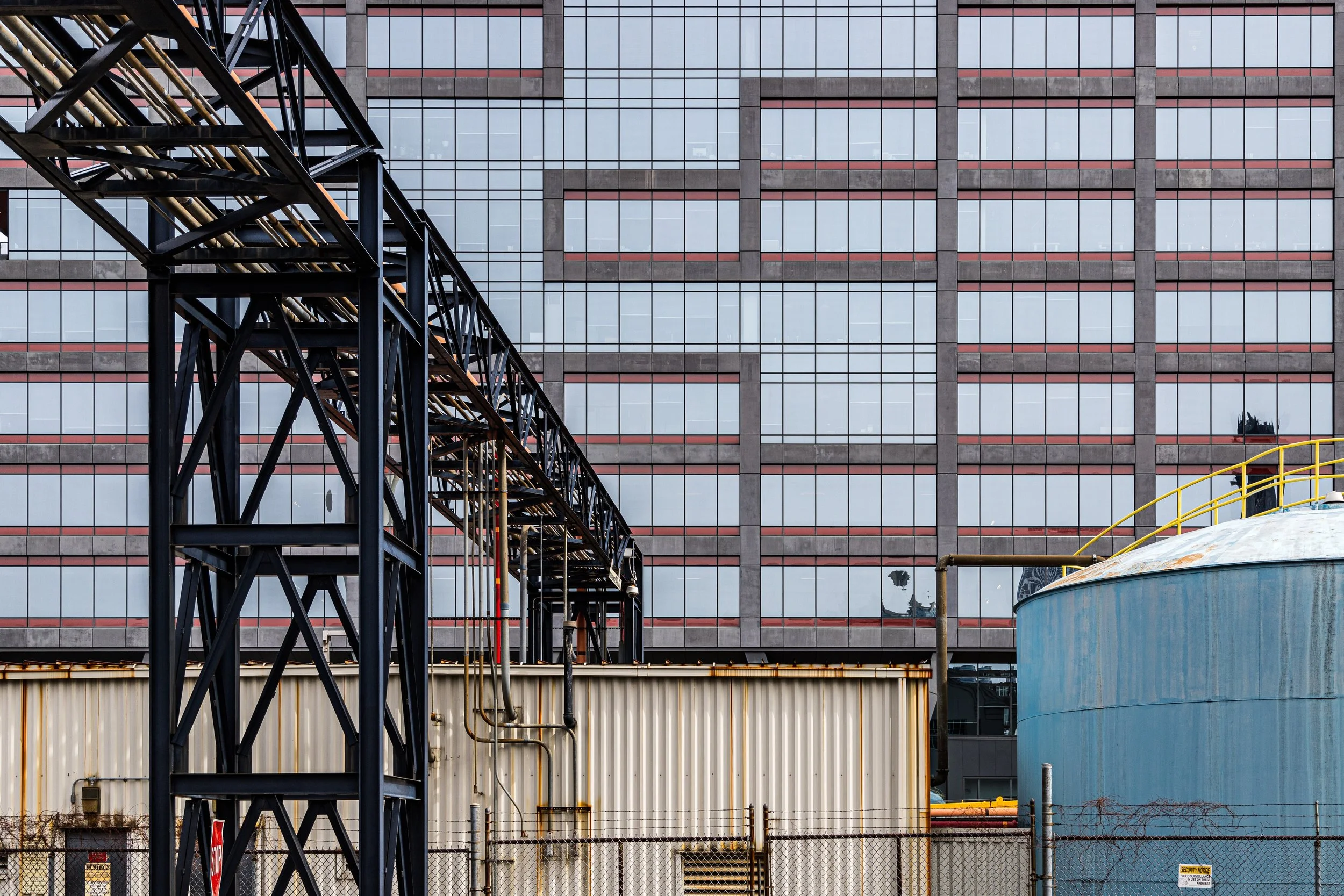 Industrial scene with a large glass building in the background, a black metal framework structure, a beige shipping container, and a blue circular storage tank with yellow railing.