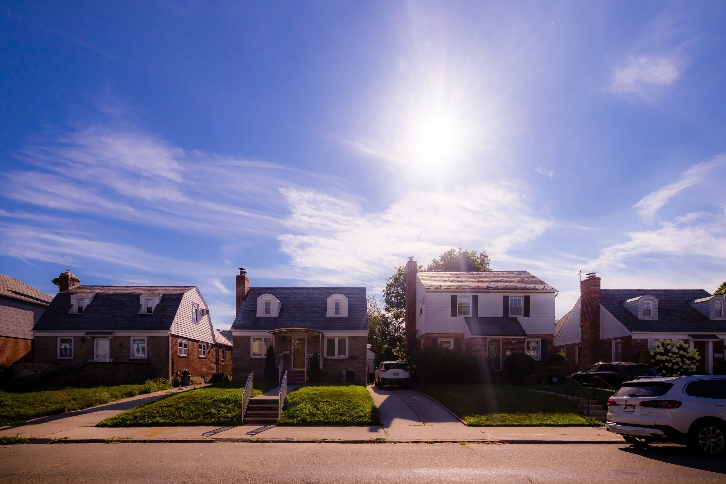 A row of three houses on a sunny day with a clear blue sky and some clouds. Cars are parked in the driveways and on the street.