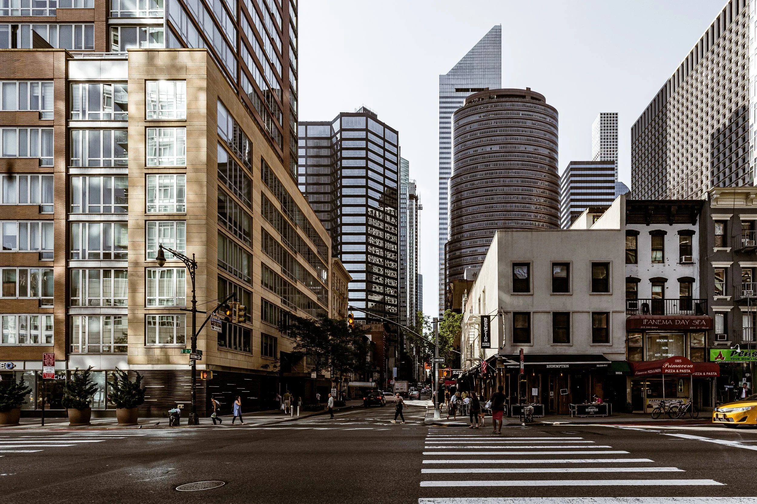 A city street scene with tall skyscrapers in the background and a mix of modern and older buildings in the foreground. Pedestrians cross the street at a crosswalk, and a few cars are visible, including a yellow taxi.