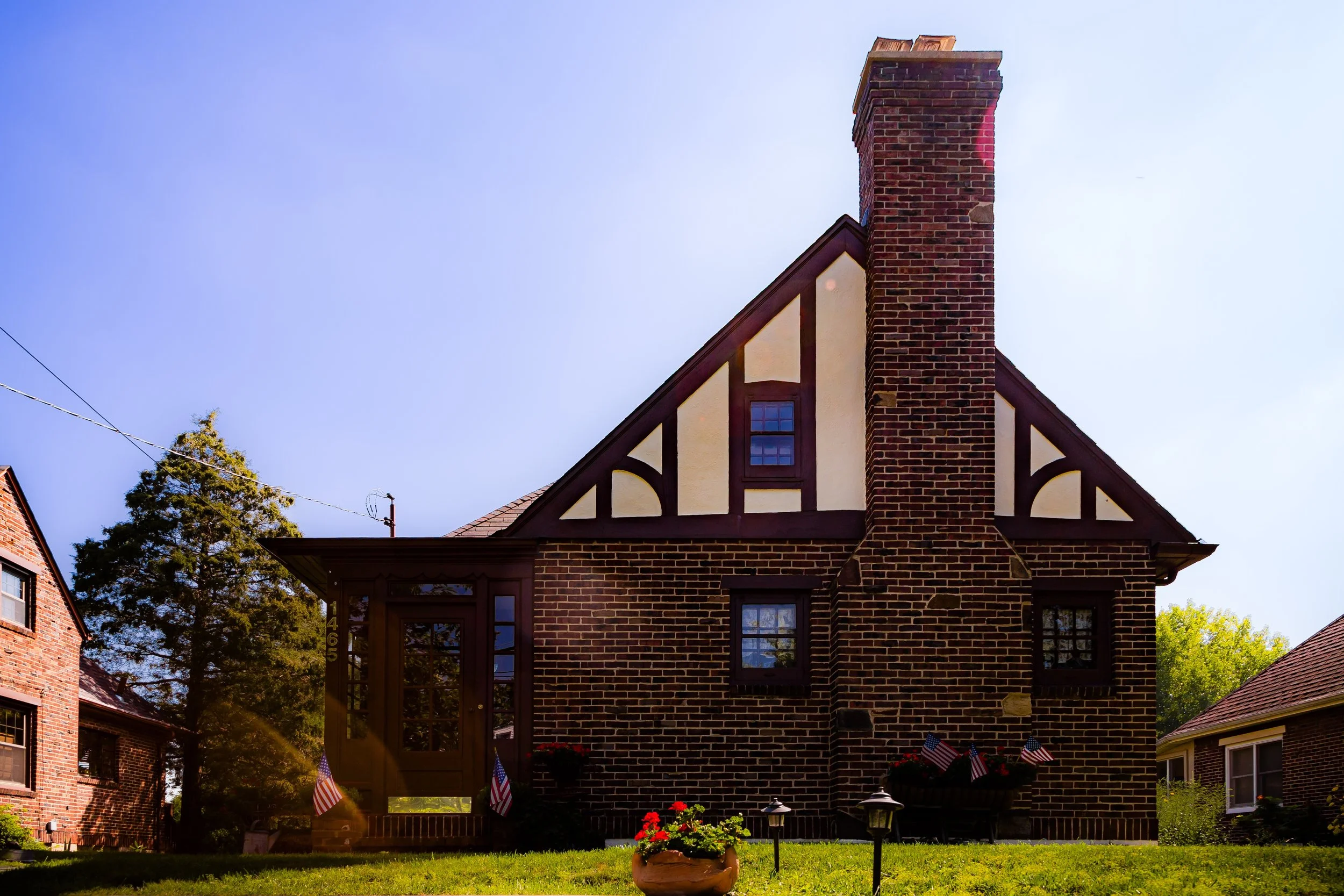 A brick house with a chimney, upper and lower windows, a glass door, and a front yard with flowers, American flags, and outdoor lights.