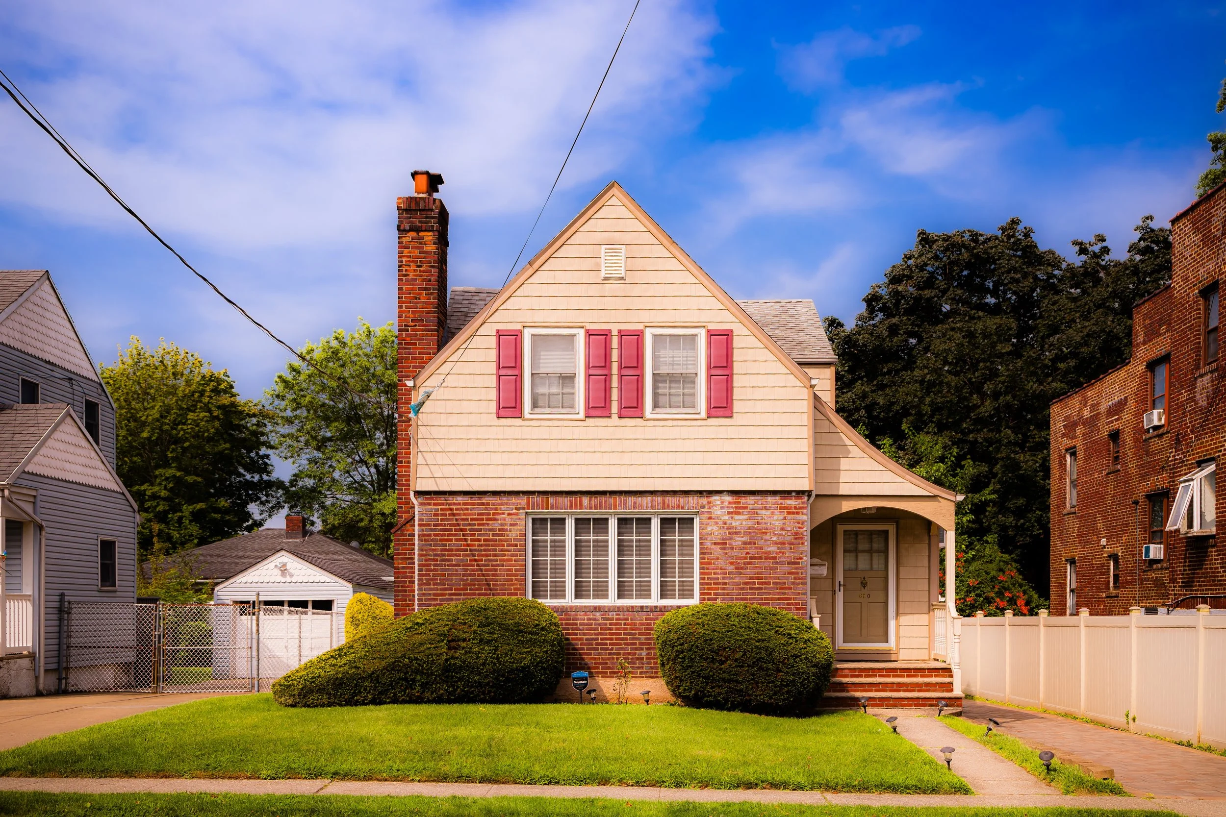 A two-story suburban house with a brick first floor, beige siding on the upper floor, and pink shutters on the second-story windows. The house has a front porch with stairs leading to the front door, surrounded by a manicured lawn and bushes. There is a white fence on the side, and neighboring houses and trees are visible in the background under a blue sky.