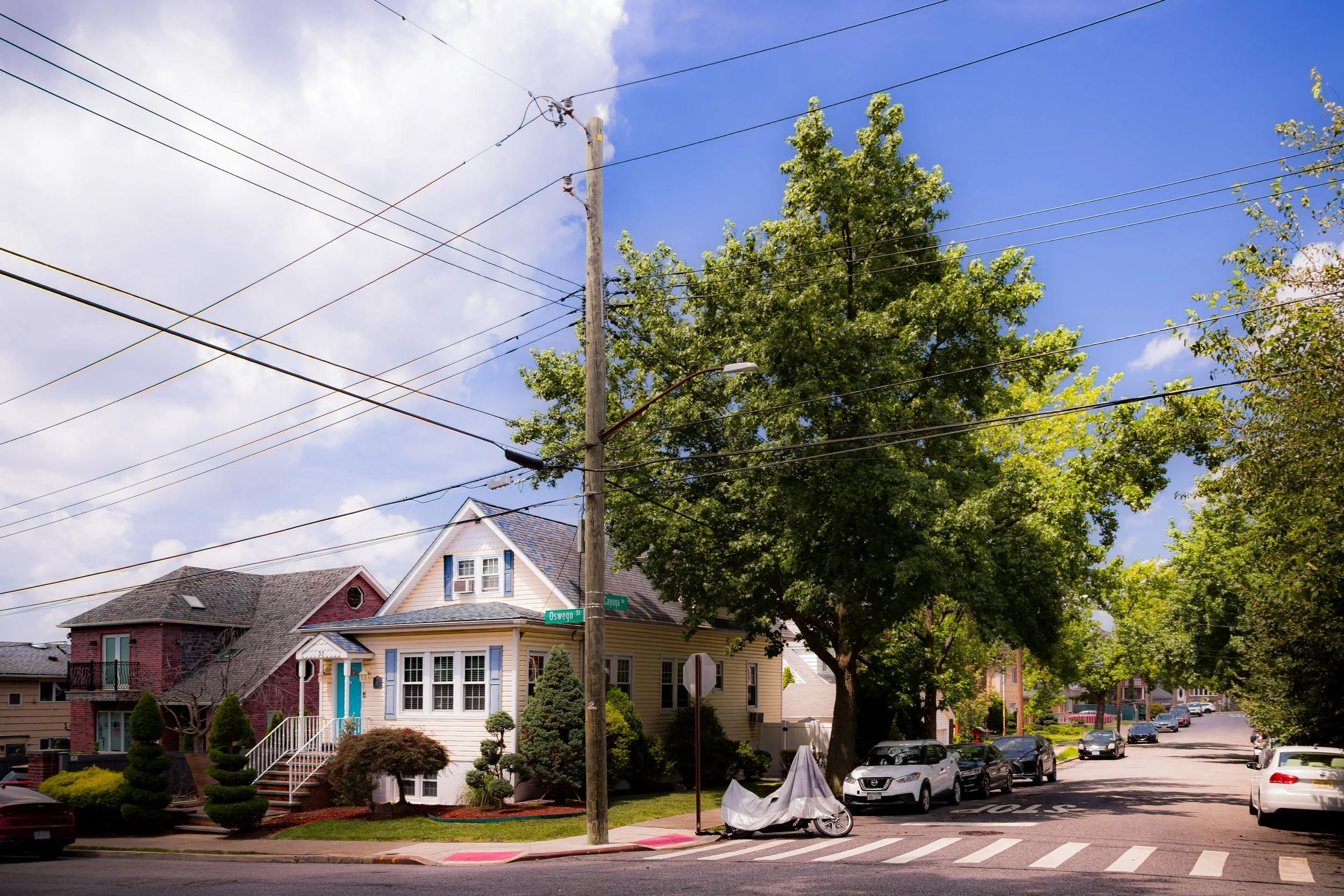 Residential street scene with houses, trees, parked cars, and a utility pole with power lines under a partly cloudy sky.