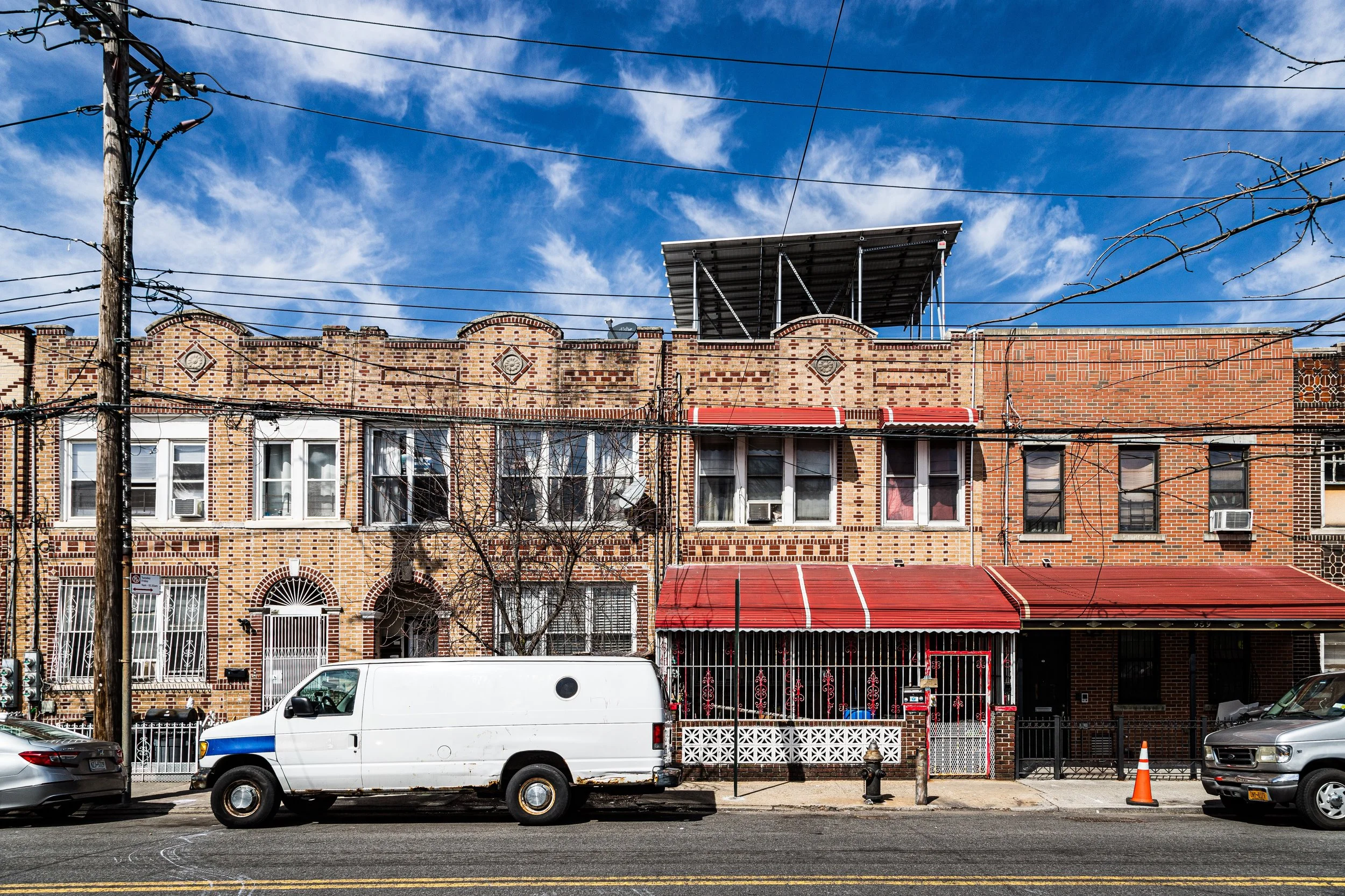 Street view of a row of brick residential buildings with red awnings, parked cars, utility poles, and electrical wires under a partly cloudy blue sky.