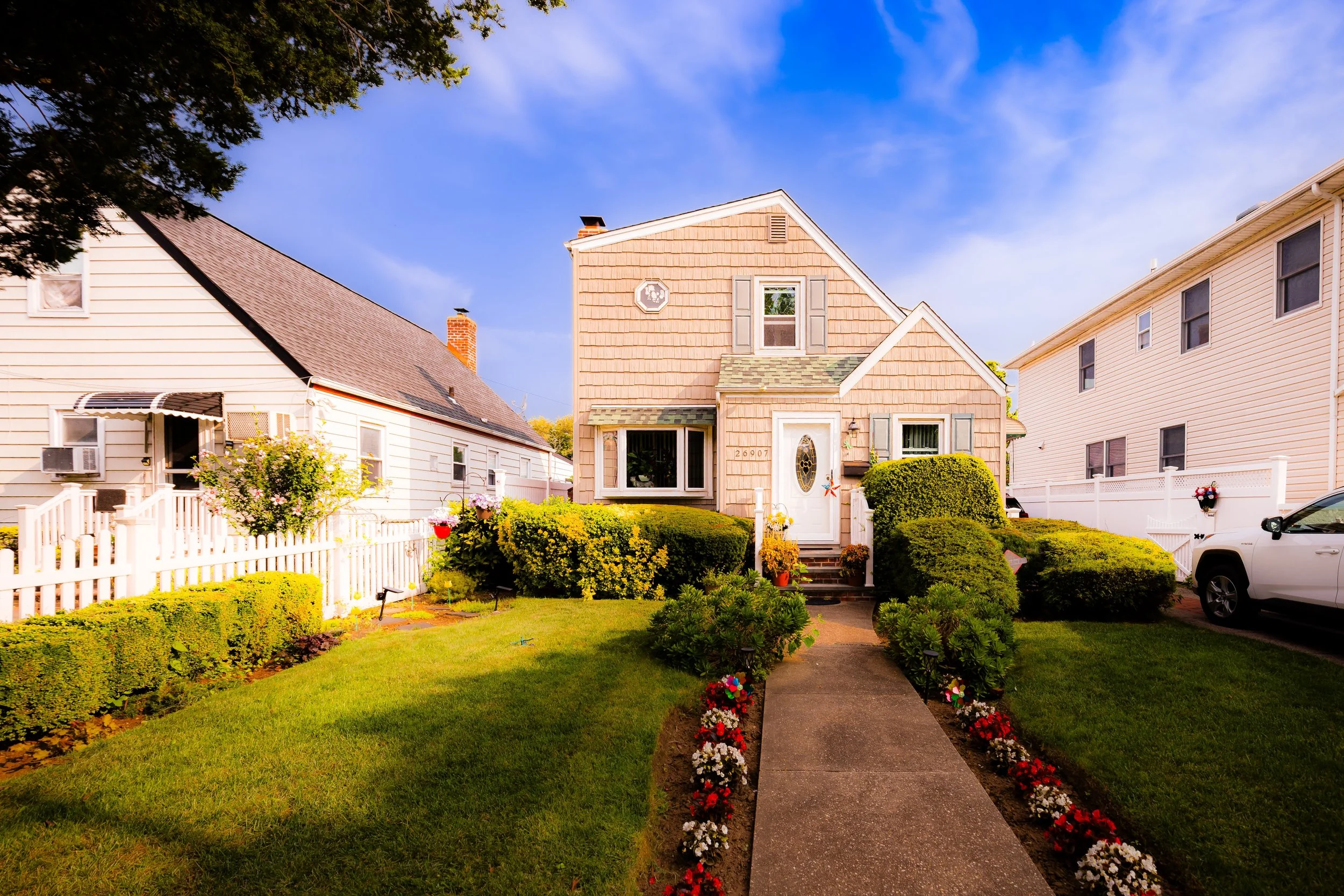 Front view of a house with a landscaped garden, a walkway lined with red and white flowers, and neighboring houses on each side under a blue sky.