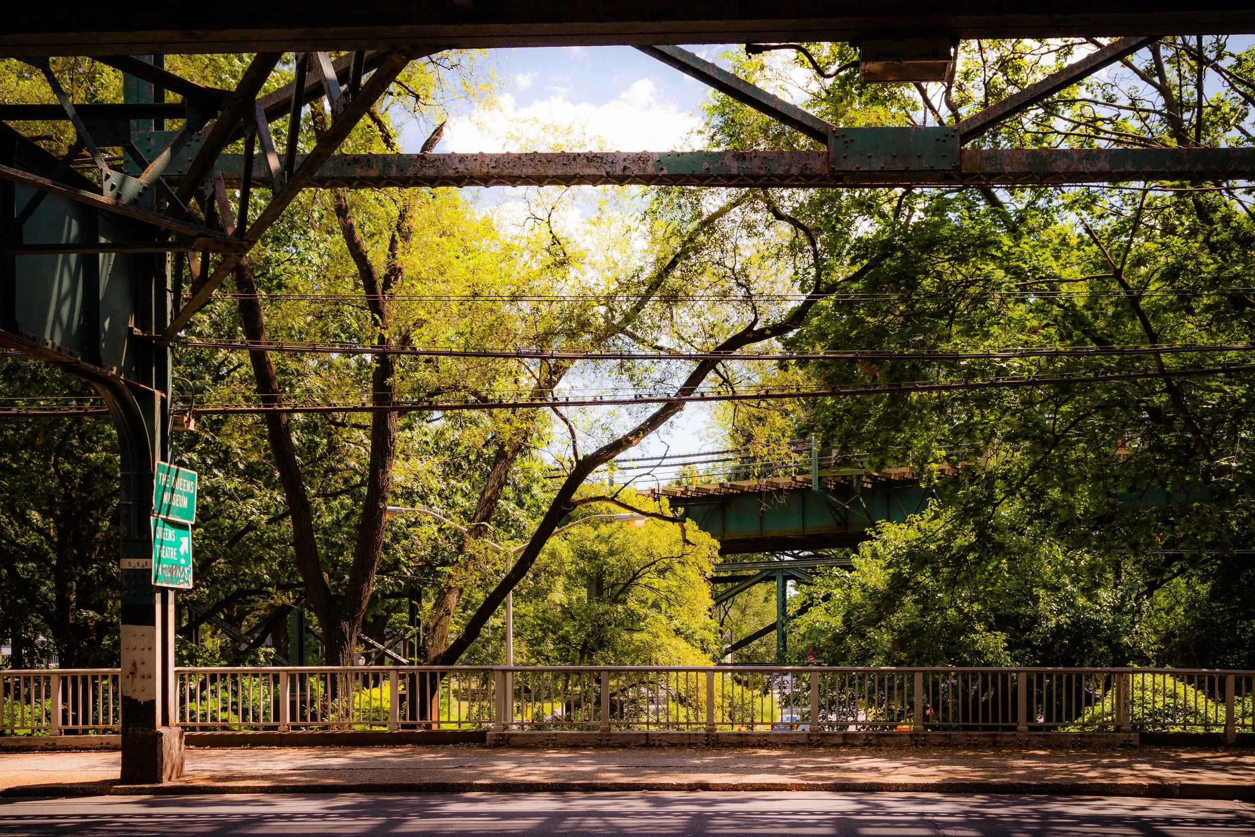 City street with trees, a metal bridge structure overhead, traffic signs, and a sidewalk with a railing.
